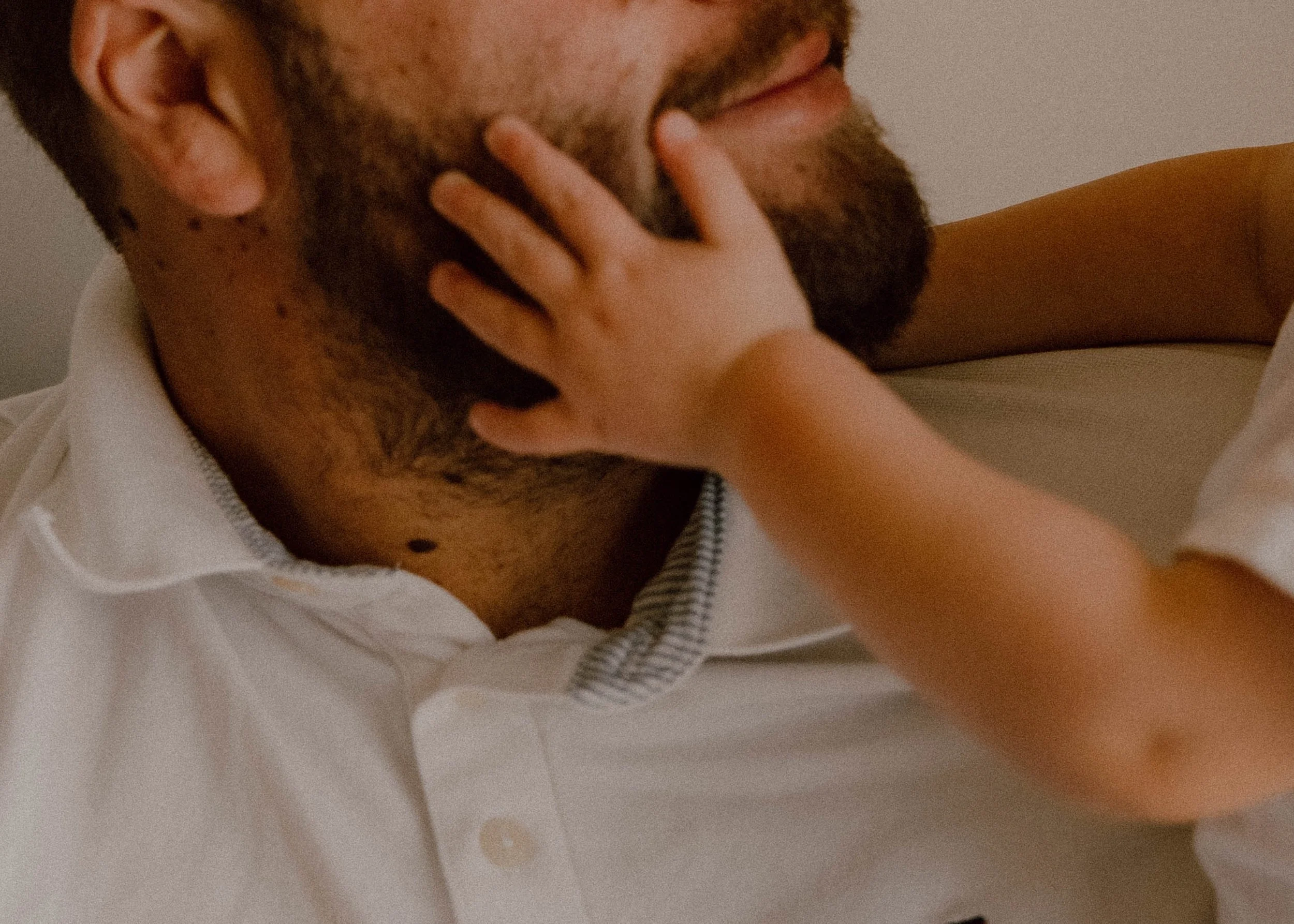 Close-up of a man with a beard touching his face with his hand, wearing a white shirt with a striped collar. candid family photos with connection as the focus