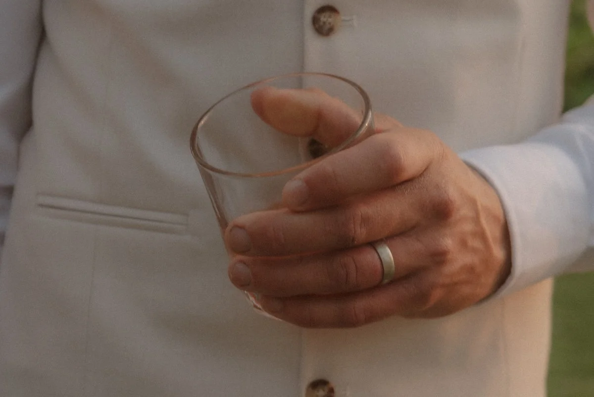 A person wearing a light gray blazer holding a clear glass of liquid in their right hand, with a silver wedding band on their ring finger. yarra valley and Melbourne wedding photographer