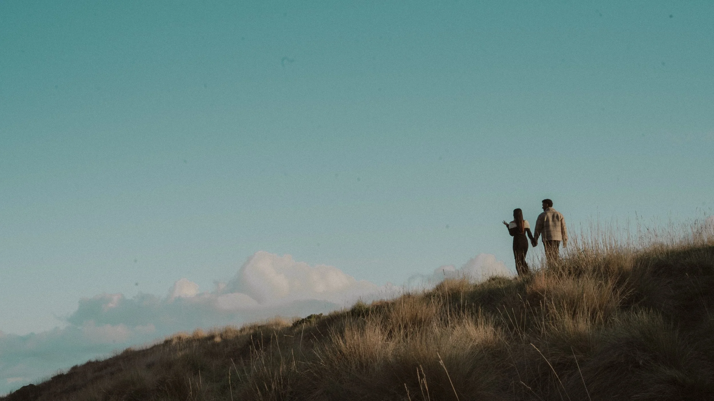 A couple walking hand-in-hand on a grassy hillside during sunset, with a clear sky and some clouds in the background. moments after their proposal/engagement with jess a yarra valley and Melbourne wedding photographer