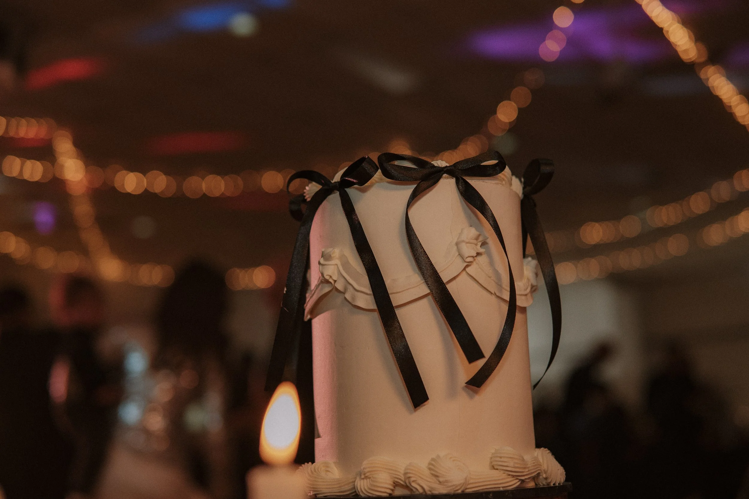 A tall white wedding cake decorated with black ribbons and white ruffles, with a lit candle in the foreground and blurred string lights in the background. yarra valley and Melbourne wedding photographer