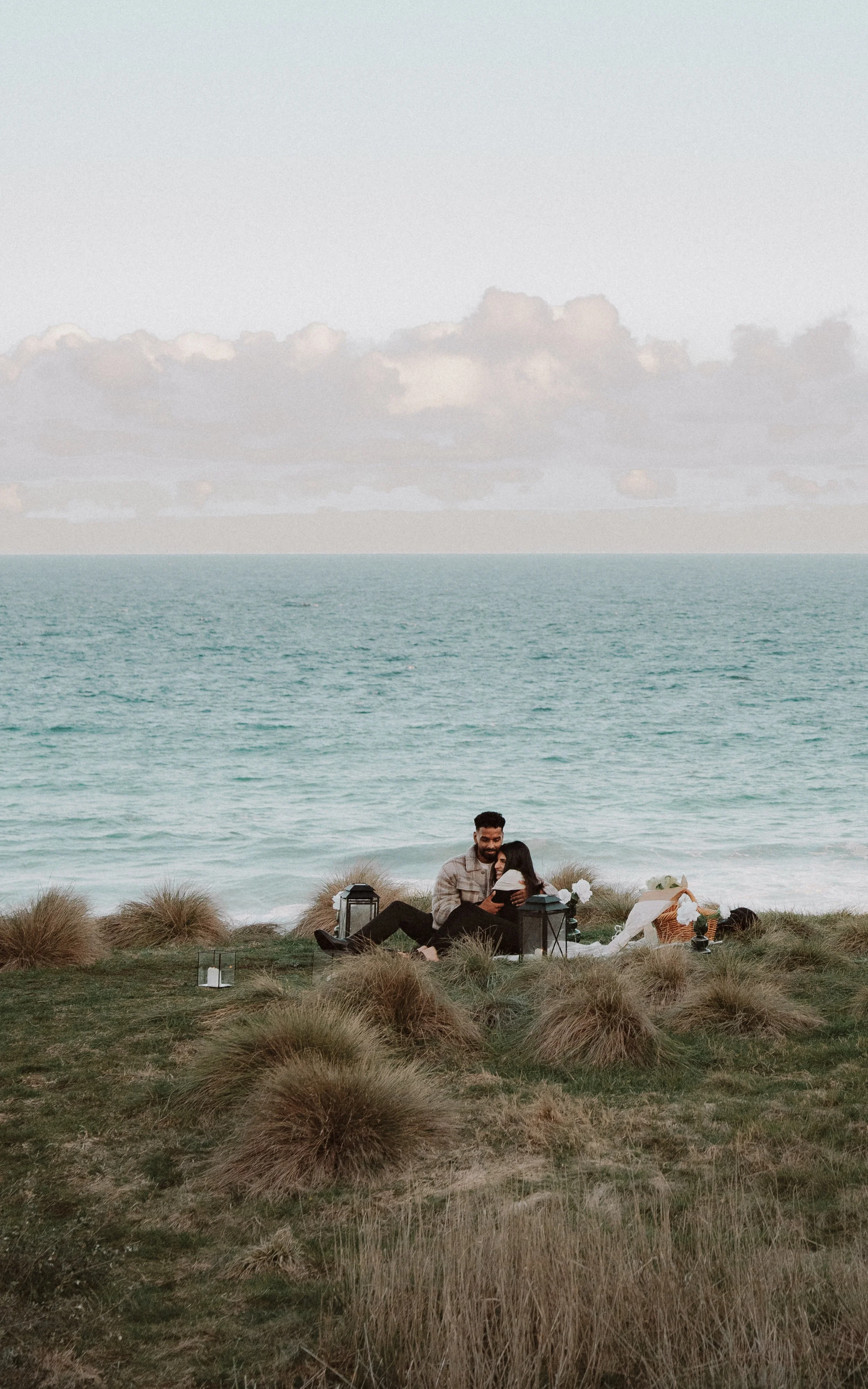 A couple enjoys a picnic on a grassy area near the ocean, surrounded by lanterns, and a floral arrangement, with the sea and cloudy sky in the background. moments after their engagement with jess a yarra valley and Melbourne wedding photographer