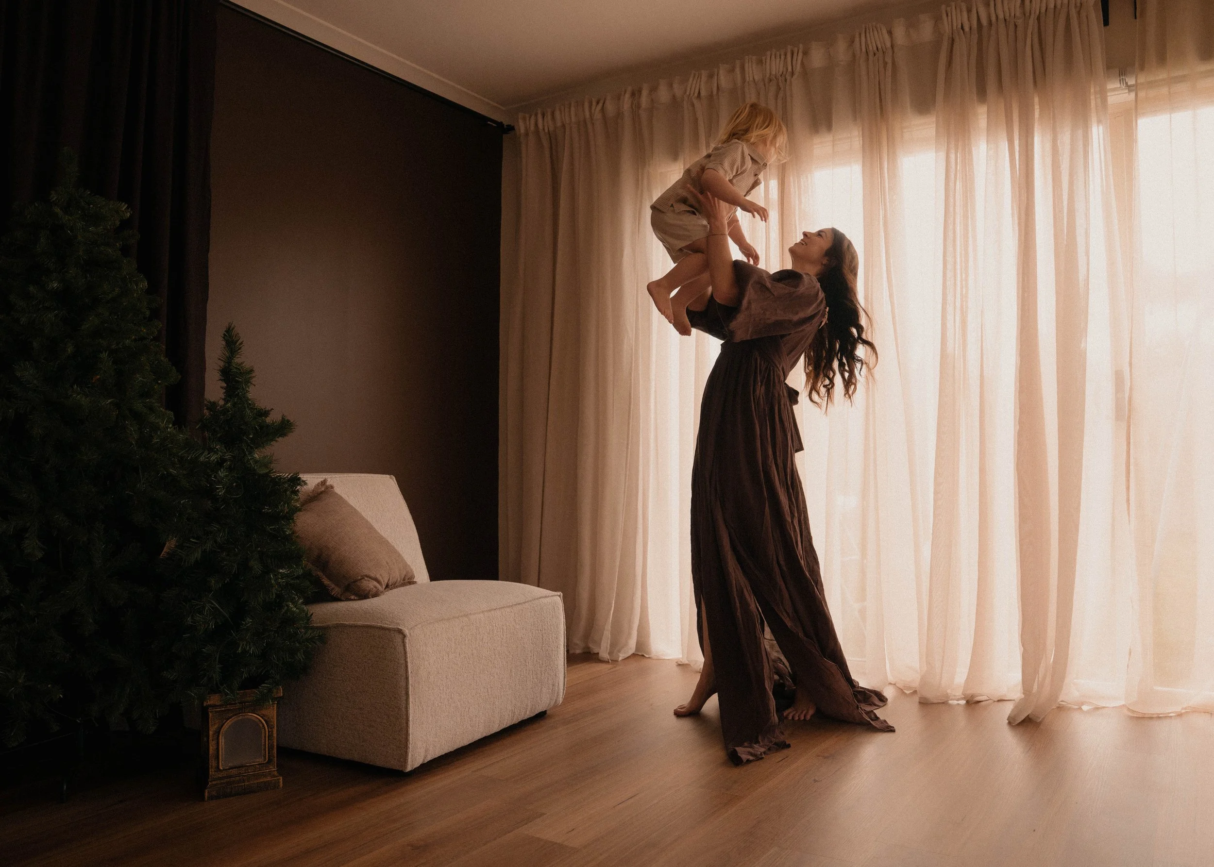 A woman lifting a young girl in the air inside a living room with beige curtains, a Christmas tree, a beige sofa, and hardwood floors.