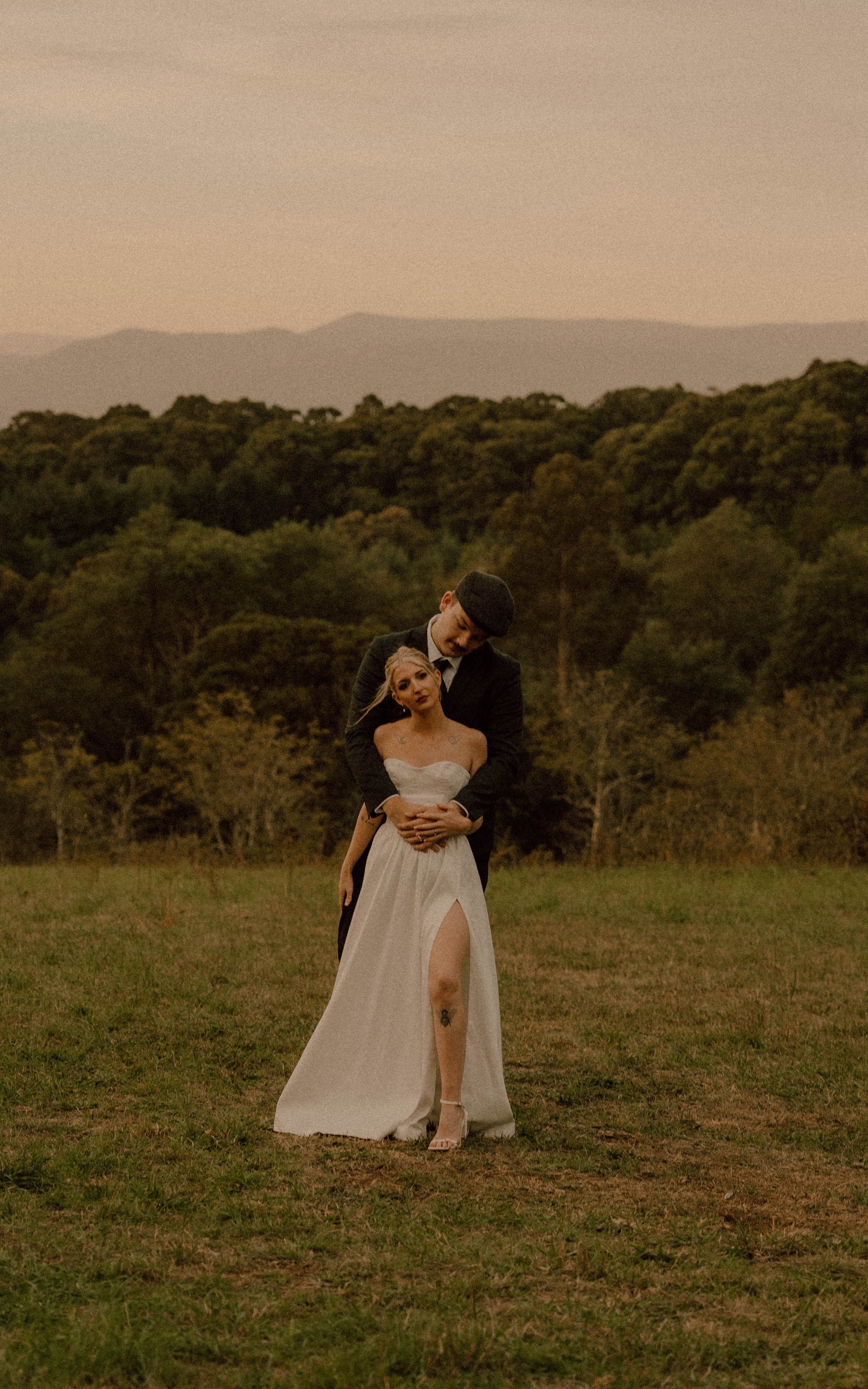 A couple dressed in wedding attire stands in a grassy field with a forested background and mountains in the distance at sunset.