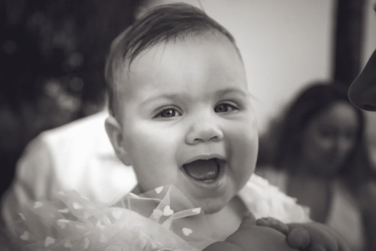 A smiling baby with short hair, looking directly at the camera, with a blurred person in the background. yarra valley and Melbourne wedding photographer