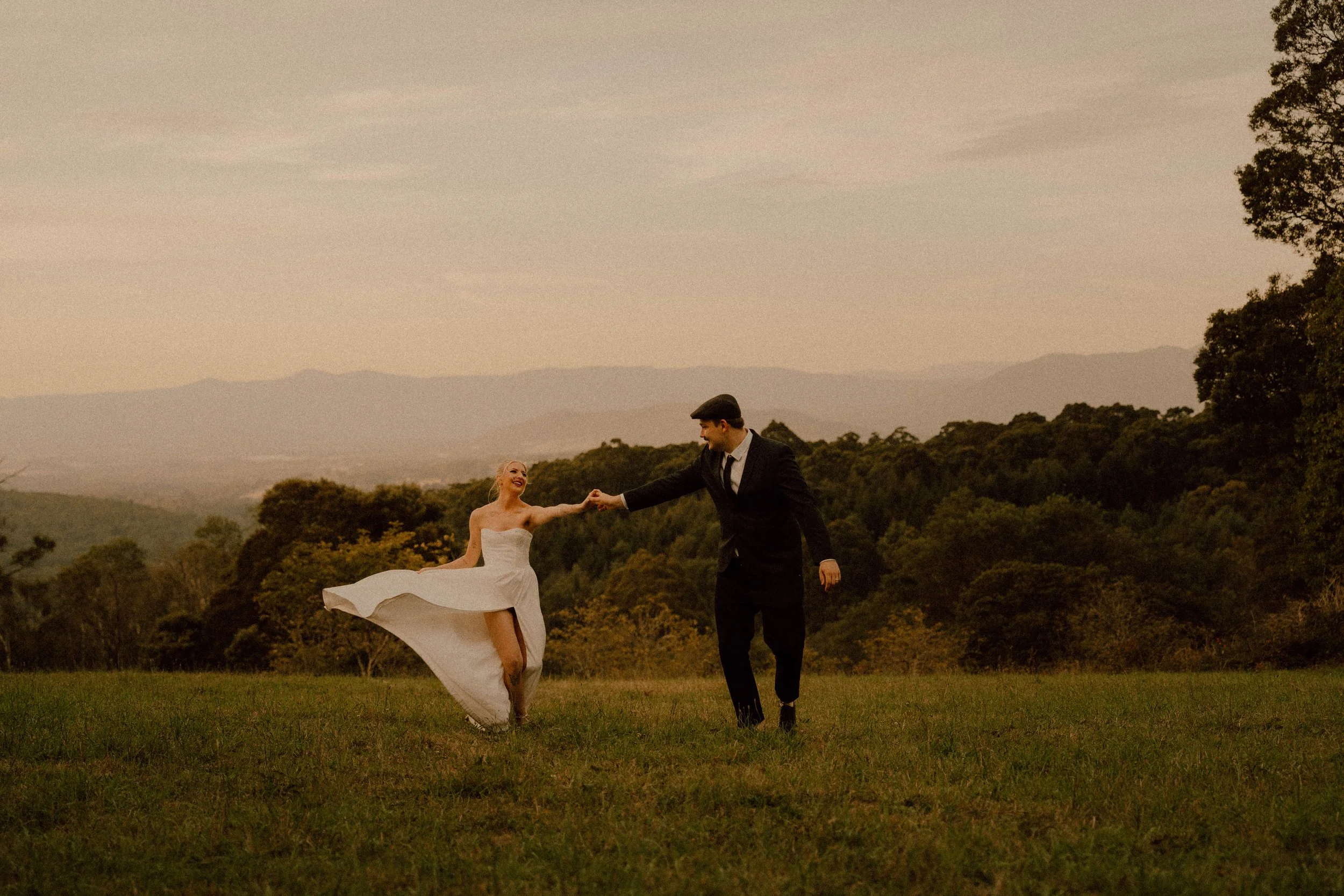 A bride and groom dancing outdoors on a field with trees and mountains in the background during sunset.