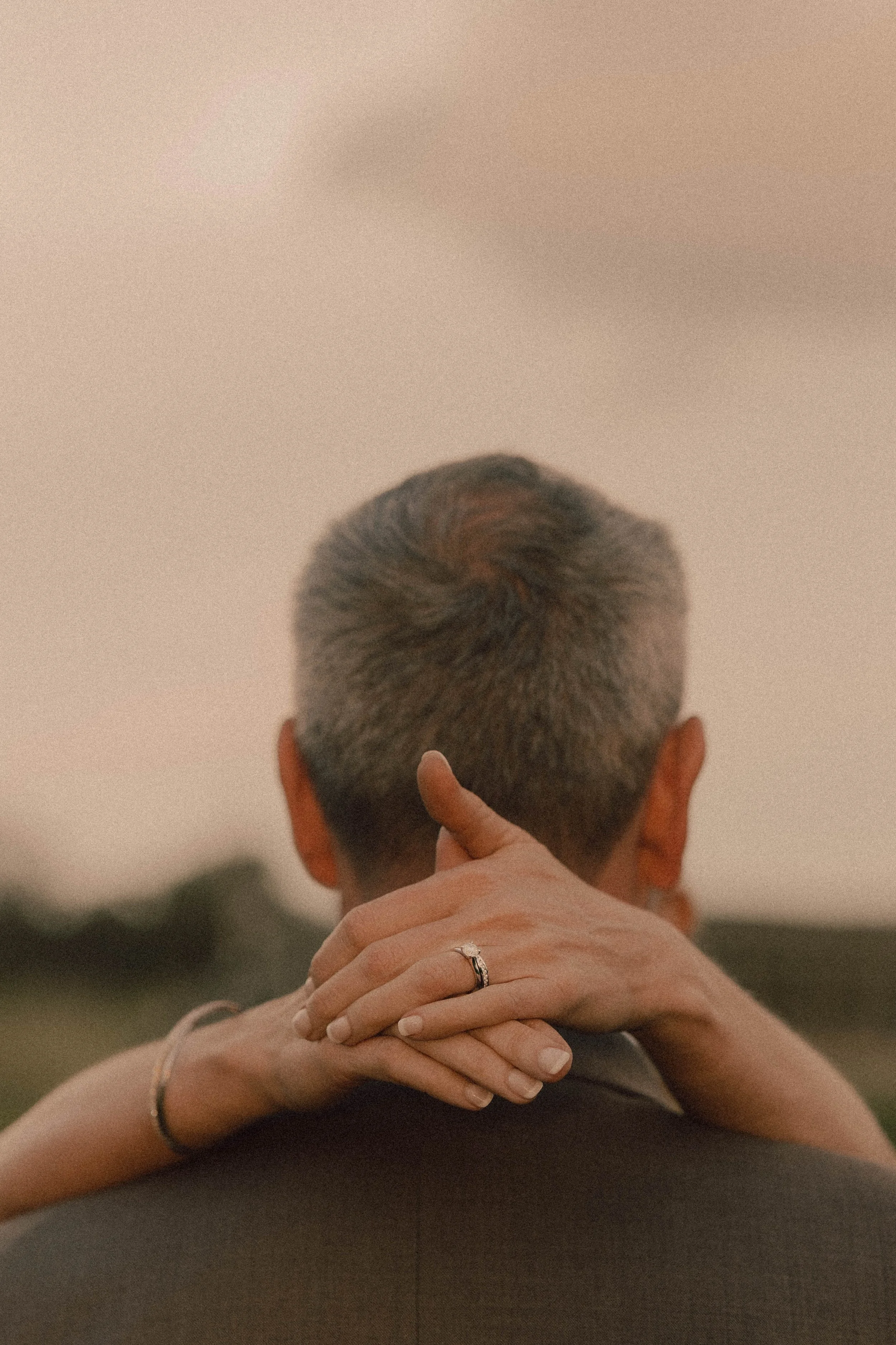 A person with short, graying hair holds their head in their hands while sitting on the ground. There is a close-up of their head from behind, and a woman’s hand with a wedding ring rests on their shoulder.