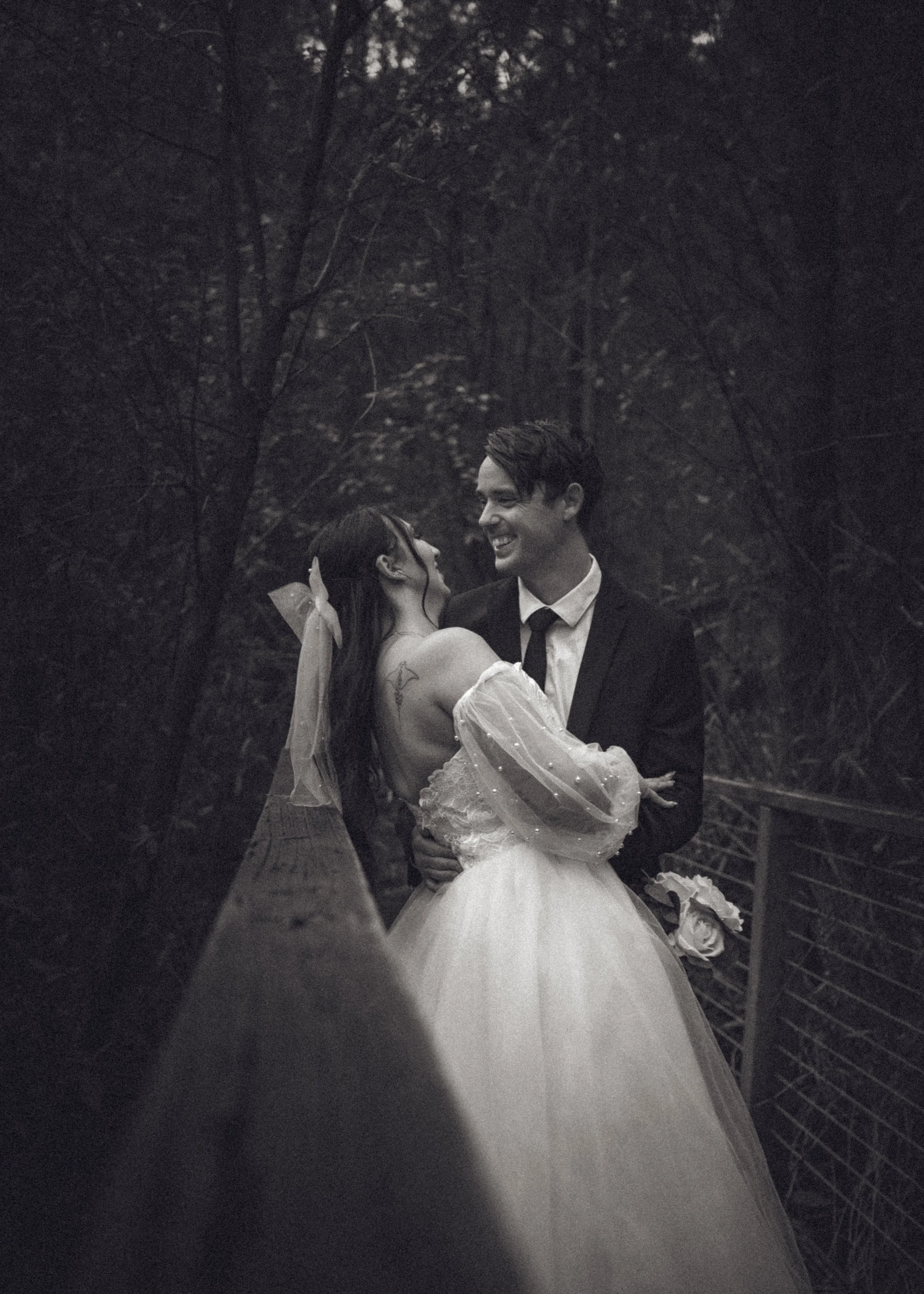 A black and white photo of a newlywed couple dancing outdoors on a bridge with trees in the background. The bride wears a dress with sheer, puffed sleeves and has a veil with a bow in her hair. The groom is in a suit and tie, smiling at the bride, ho