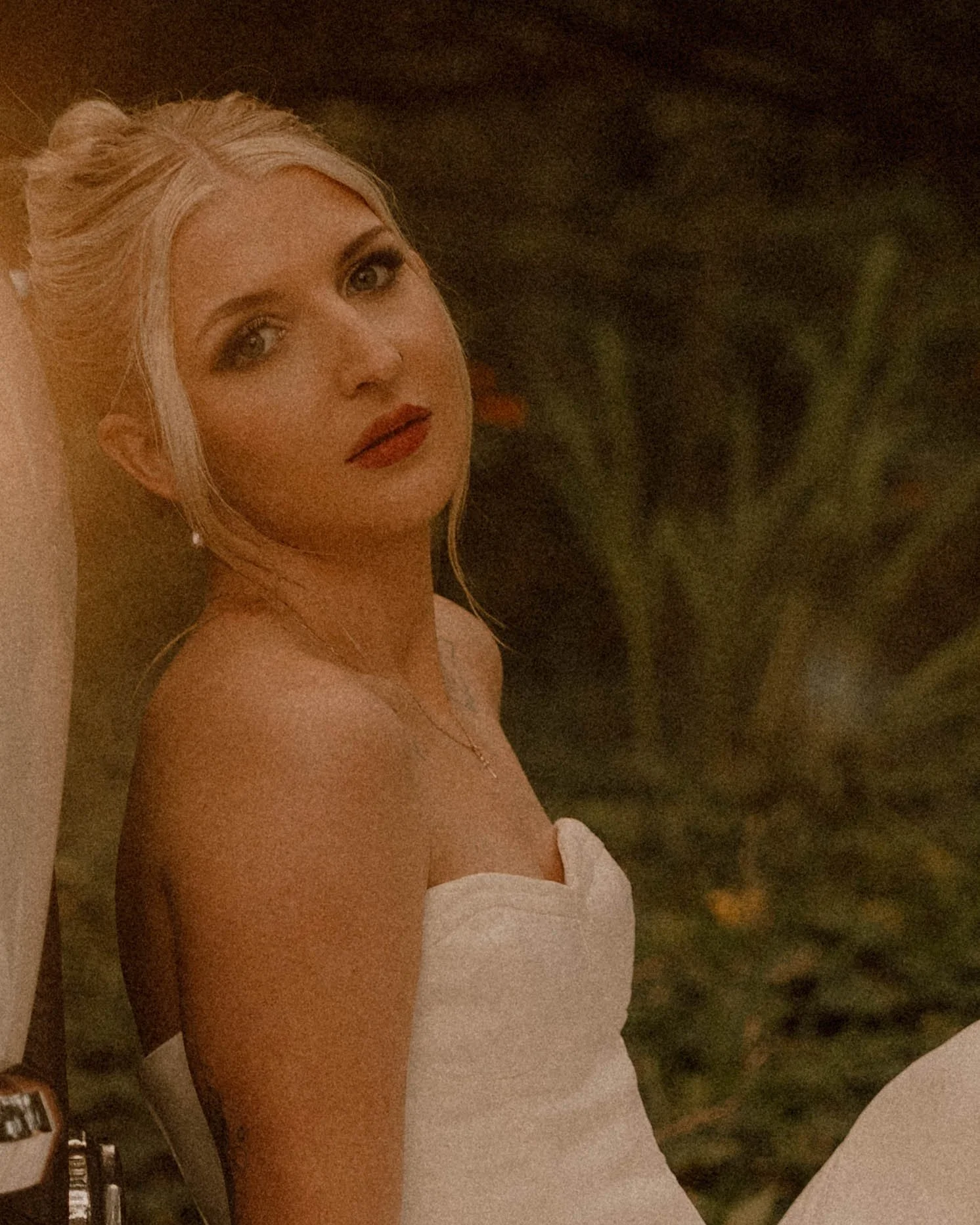 A woman with blonde hair and makeup, wearing a white strapless dress from show, is sitting  near a dark, blurred background. steaming bridal portraits