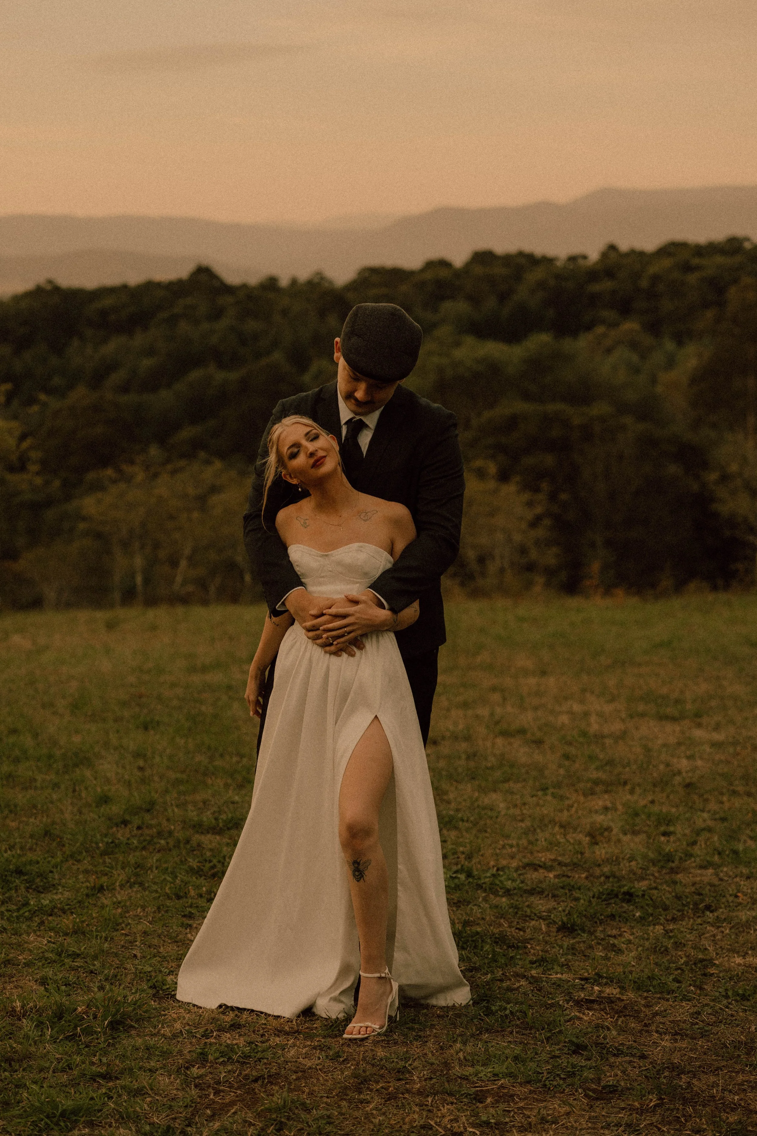 A couple dressed in wedding attire, outdoor setting with grassy field and hills in the background, woman with blonde hair in a white strapless wedding dress; man in a black suit and hat, embracing the woman from behind during sunset.