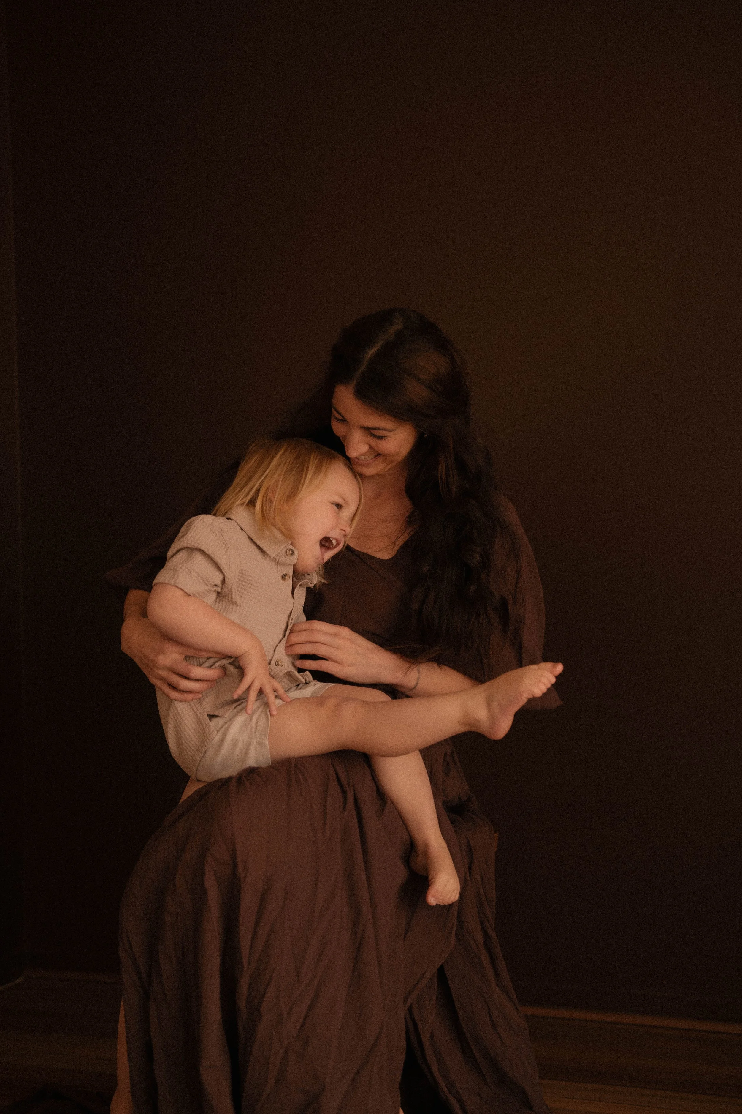 A woman with long dark hair holding a laughing little boy with blonde hair against a dark  chocolate brown background. motherhood session of a newly single mum starting over and celebrating the love for her son