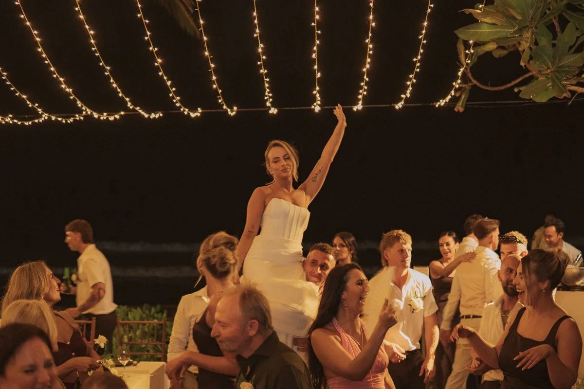 A woman in a white dress is lifted on someone's shoulders at a nighttime celebration on a beach with string lights overhead. People around her are talking and laughing, and some are holding drinks. yarra valley and Melbourne wedding photographer