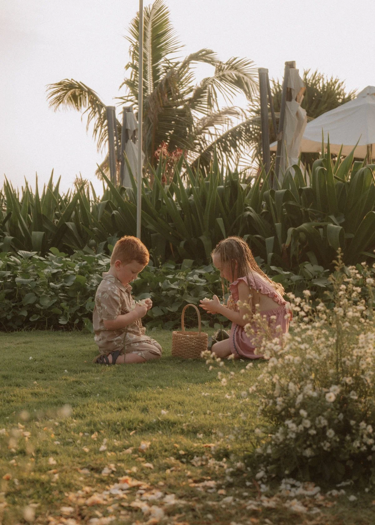 Two young children, a boy and a girl, are sitting on the grass and playing with small objects. The setting appears to be a garden or park during late afternoon or early evening, with lush greenery, large plants, and a white umbrella in the background