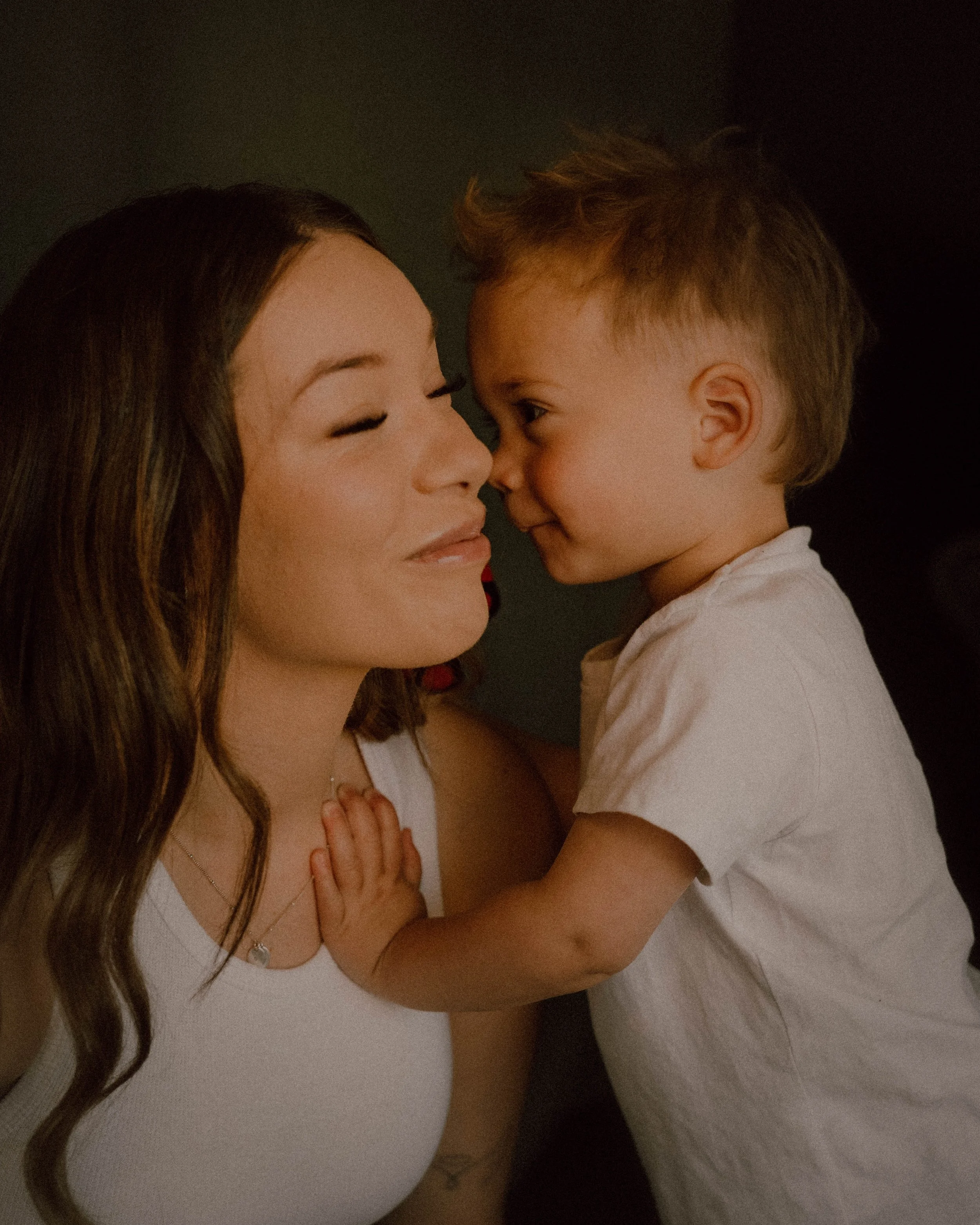 A woman with long brown hair and a young boy with short, light brown hair are touching foreheads and smiling in front of a dark background.
