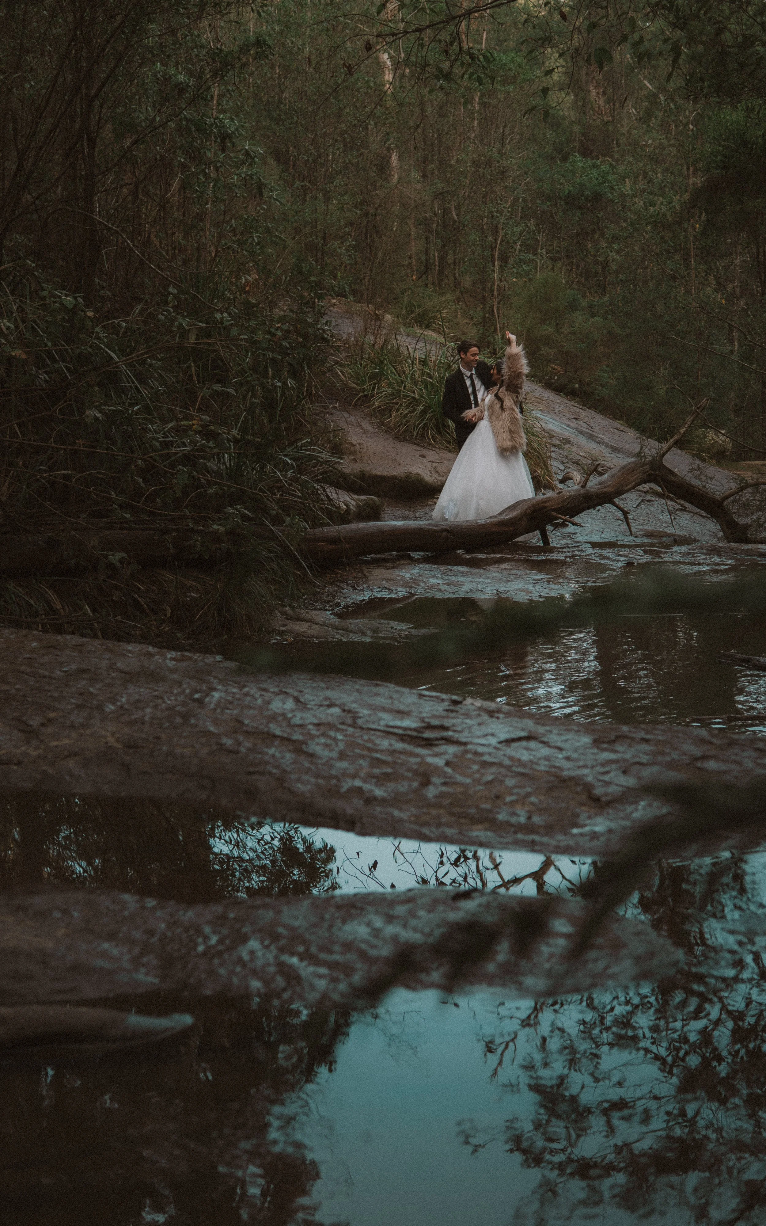 A bride and groom stand on a rocky riverbank in a wooded area, with the bride dressed in a white gown and fur stole, and the groom in a black suit, during a wedding photo shoot.