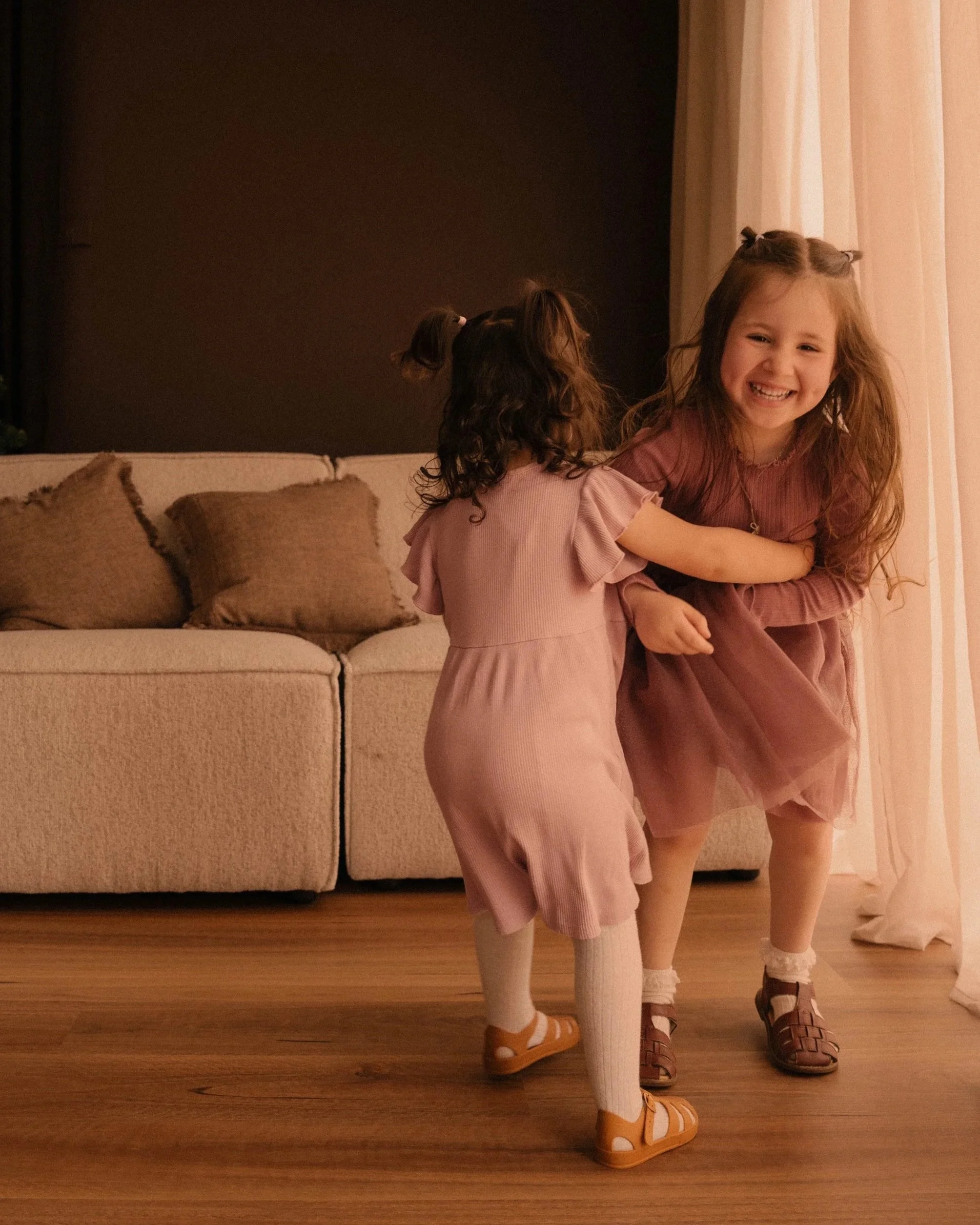 Two young girls with long hair, one with curly hair and the other with straight hair, are playing and hugging each other in a cozy living room, dressed in pastel-colored outfits, with a beige sofa and curtains in the background.