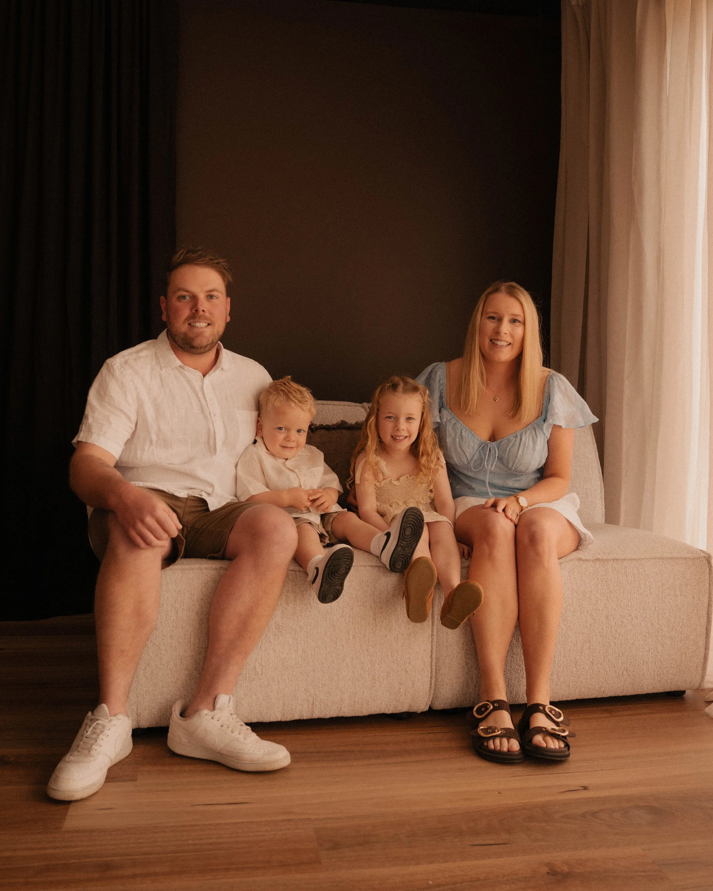 Family of six sitting on a beige couch in a room with dark curtains on the left and light curtains on the right, smiling and posing for a photo.
