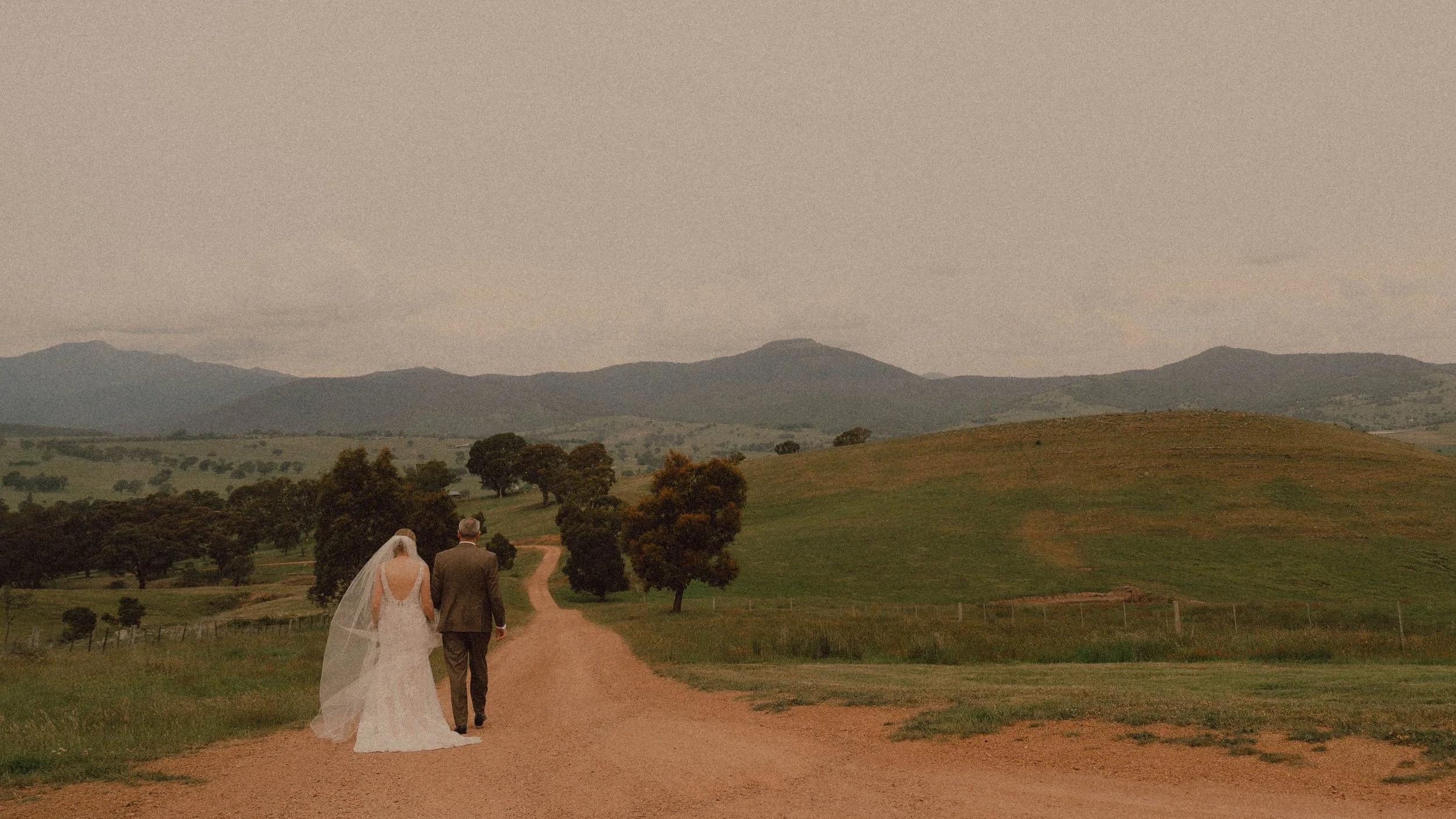 A bride and groom walking on a dirt path through a scenic green landscape with trees and rolling hills in the background, mountains in the distance, and a cloudy sky overhead.