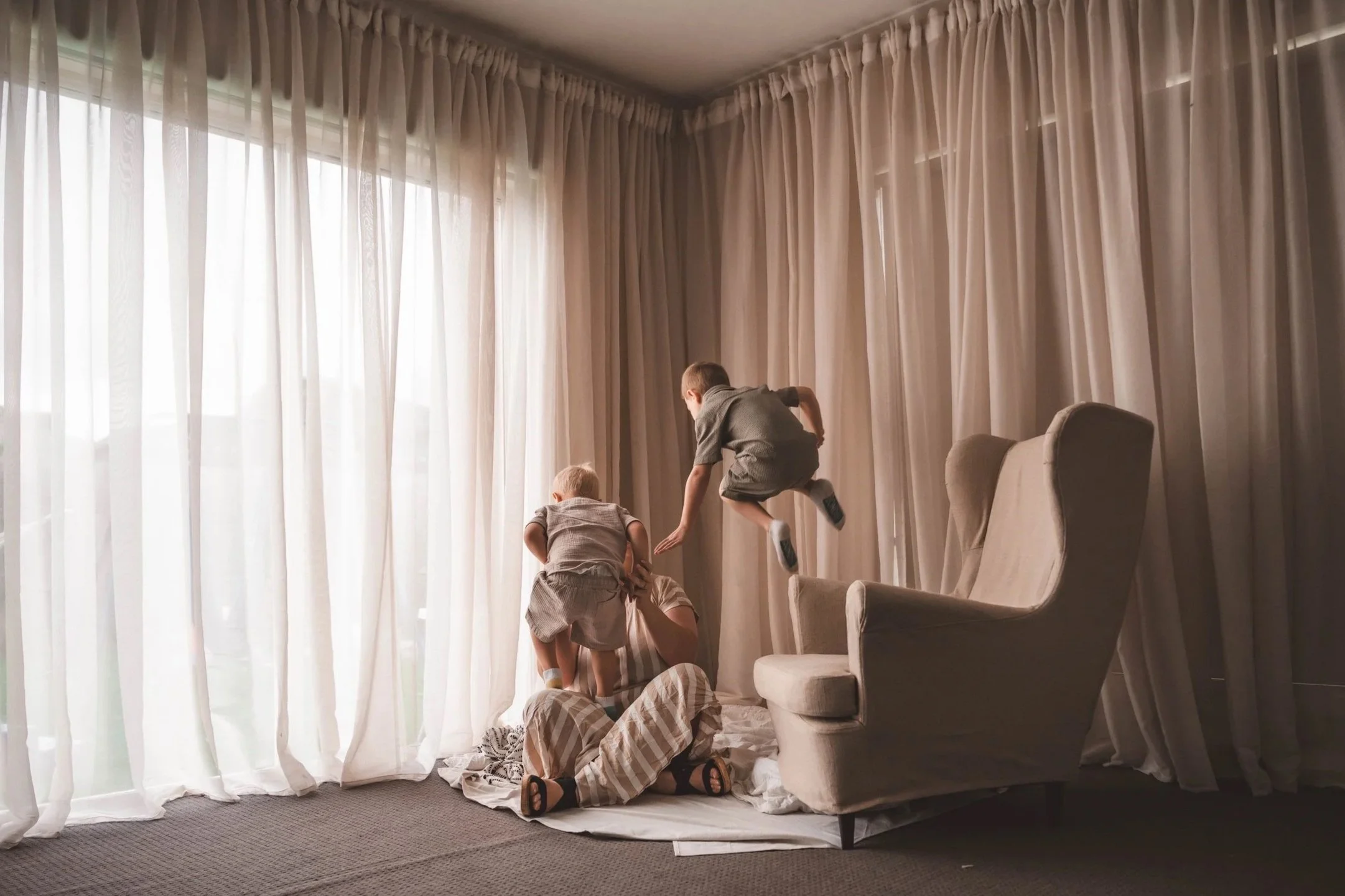 Two children and an adult playing on a bed in front of floor-to-ceiling curtains, with the children jumping and the adult sitting on the bed.