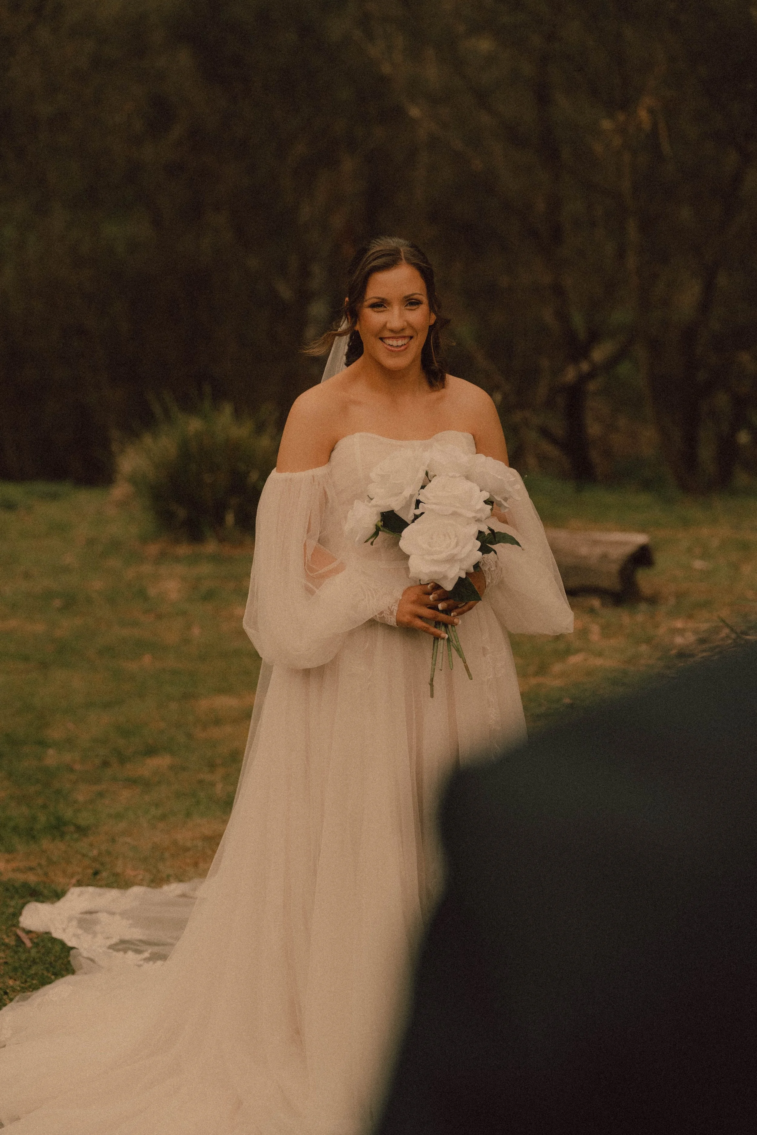A smiling bride holding a bouquet of white roses outdoors, wearing an off-shoulder wedding dress, with a background of trees and park benches.