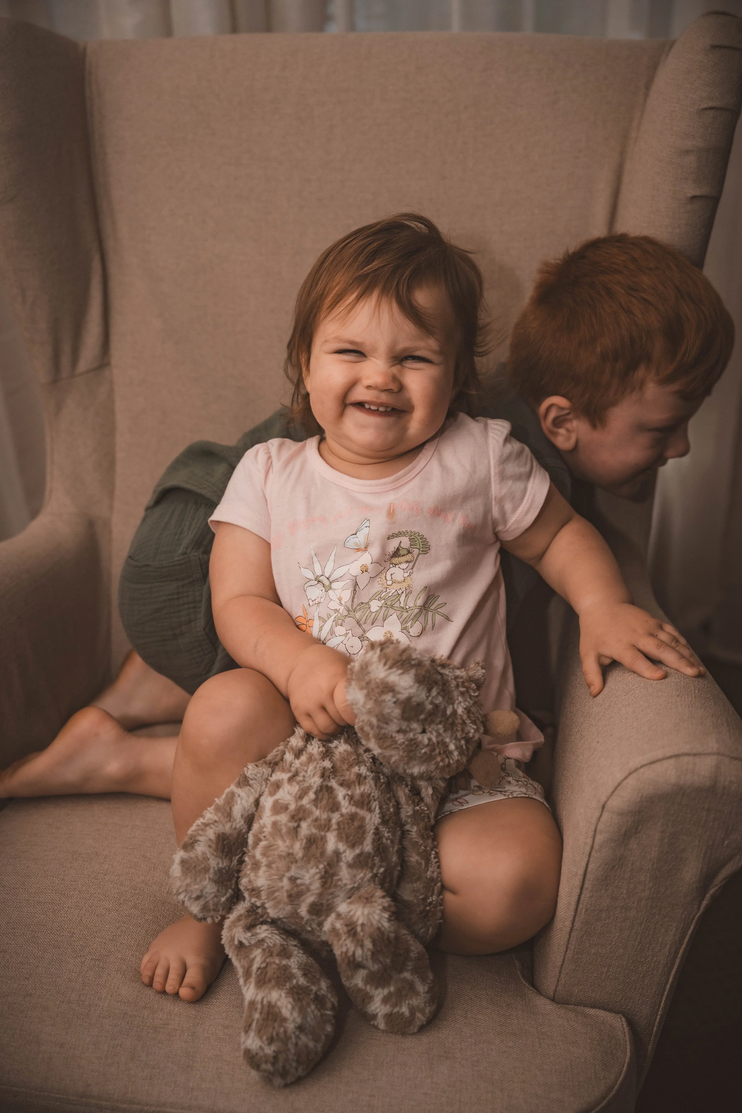 A young girl with a big smile sitting on a beige armchair, holding a plush giraffe toy. She has short brown hair and is wearing a pink shirt with a floral and butterfly design. A boy with short red hair is behind her, leaning over the armrest, facing away from the camera.