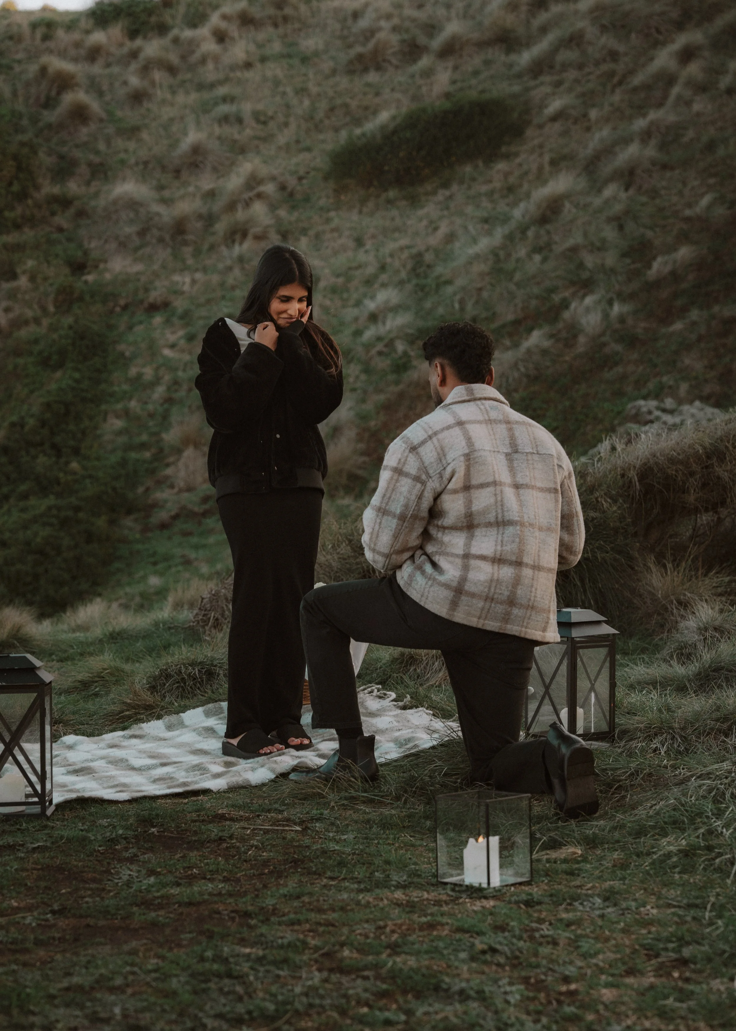 A couple is having an outdoor romantic proposal at sunset on a grassy hillside with lanterns and candles around. moments after their proposal/engagement with jess a yarra valley and Melbourne wedding photographer