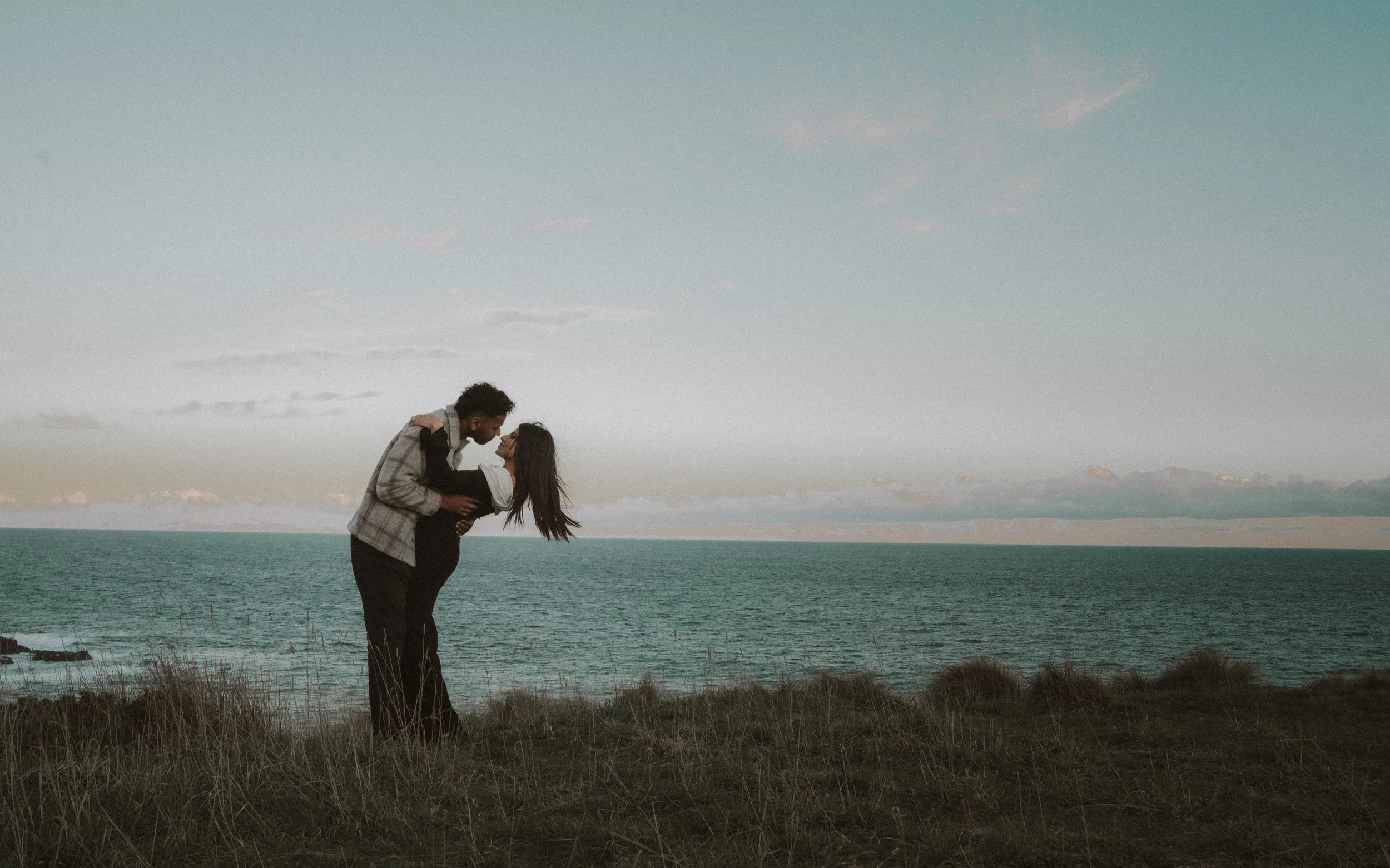 A couple at the beach, with the man dipping the woman backward, both smiling, standing on grass with the ocean and sky in the background. moments after their proposal/engagement with jess a yarra valley and Melbourne wedding photographer