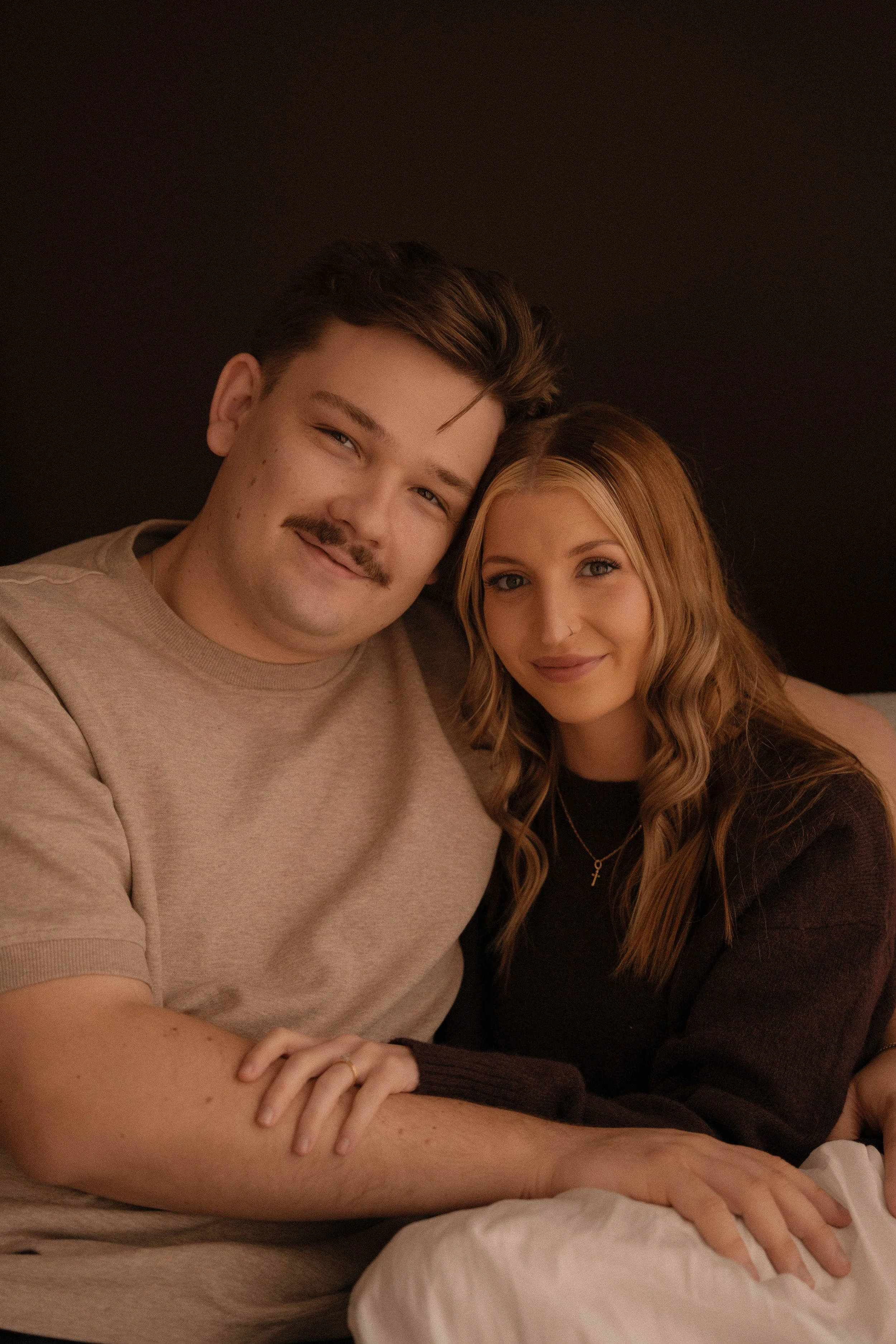 A young man with a mustache and dark hair smiling and a young woman with blonde hair and a nose piercing smiling, sitting close together with their arms overlapping, against a dark background.