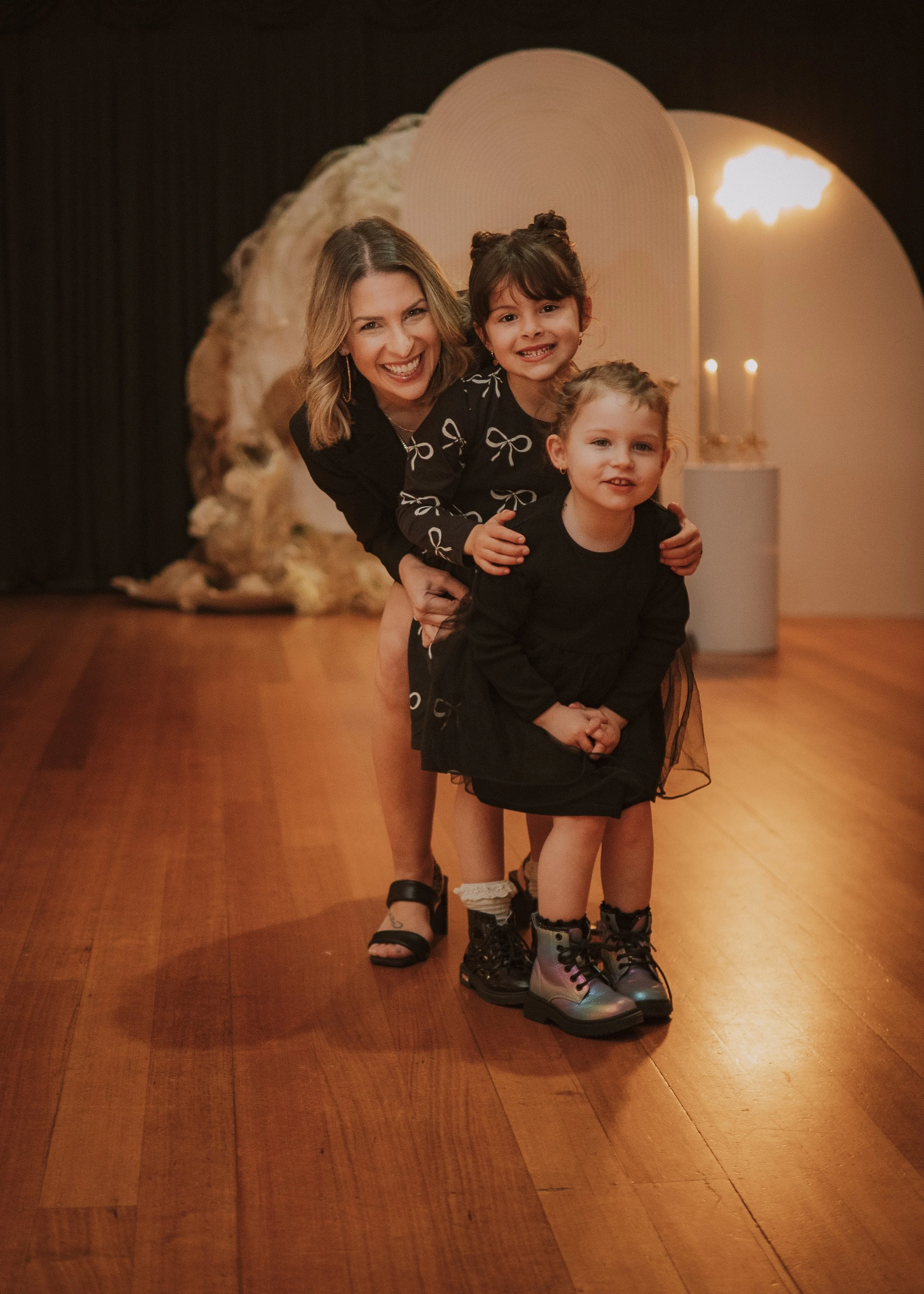 A woman and two young girls smiling and posing together indoors with warm lighting, wooden floor, and decorative background elements. yarra valley and Melbourne wedding photographer