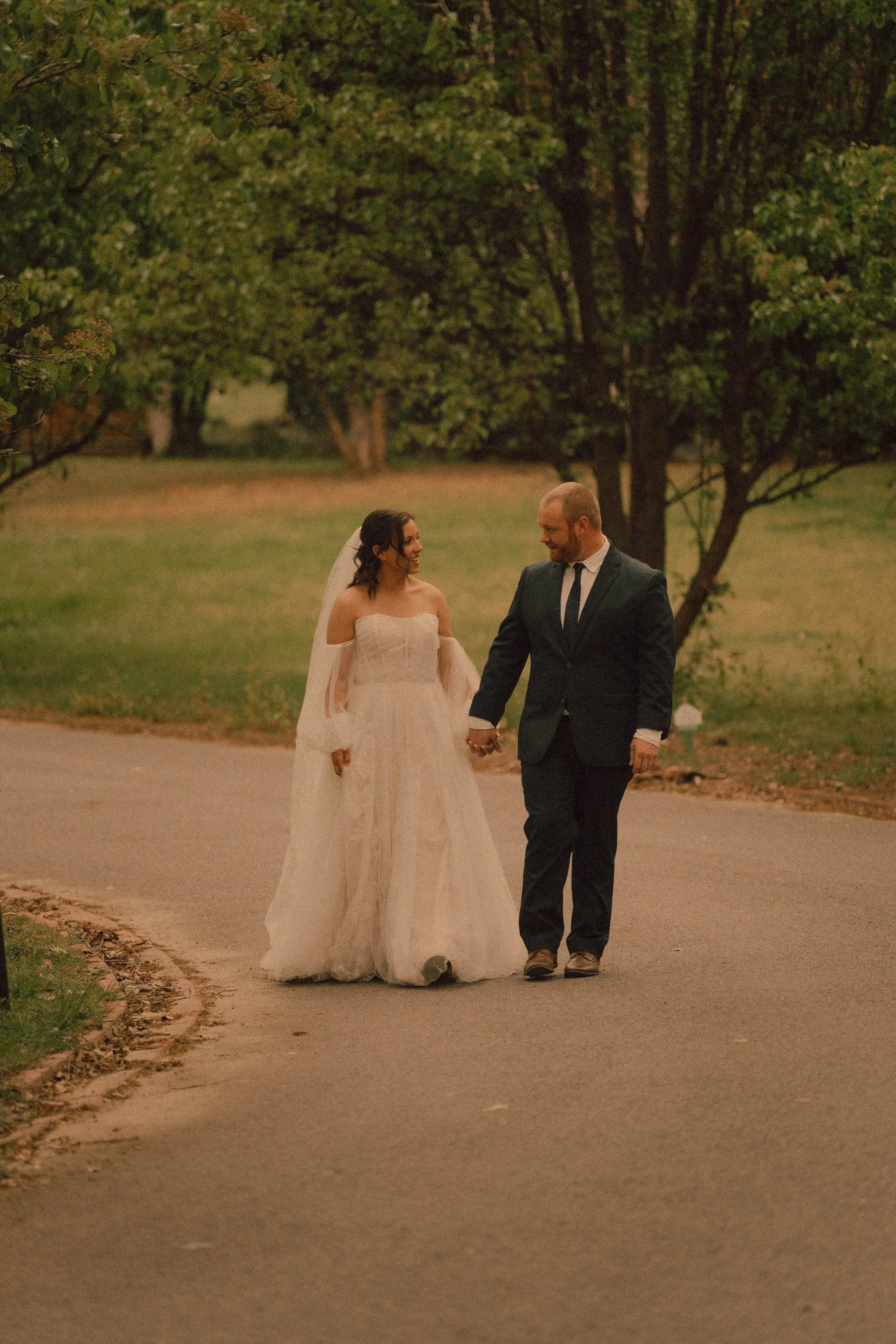 A bride and groom walk hand in hand outdoors, smiling at each other, during a twilight moment in a park with green trees.