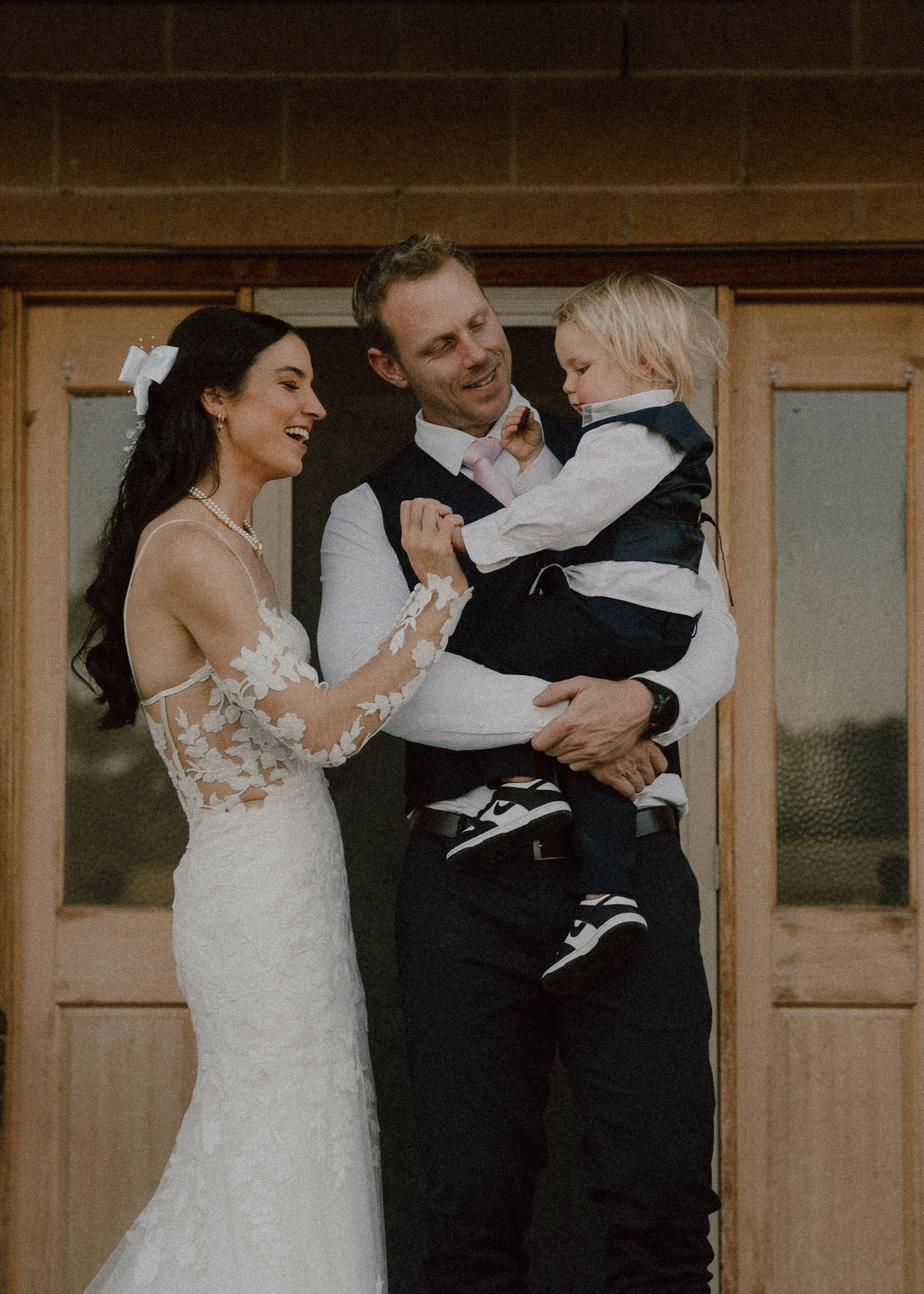 A bride in a wedding dress from brides of Werribee and veil smiling, groom in a shirt, tie, and vest holding a young boy dressed in a shirt, vest, and striped pants at a wedding ceremony. the little family celebrating their love