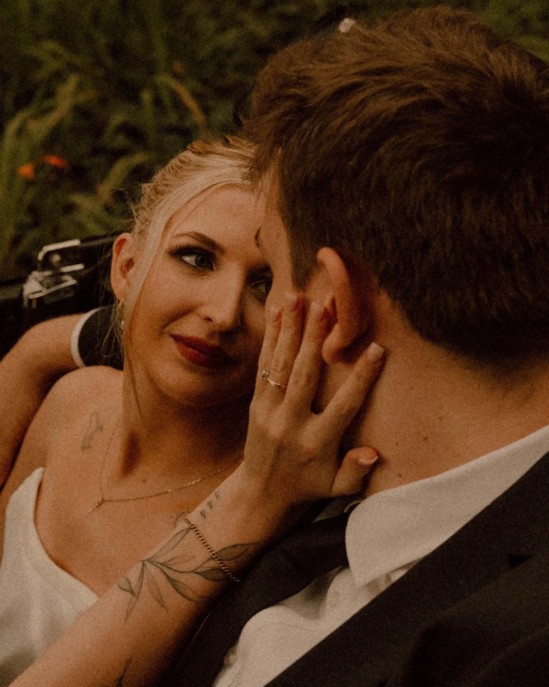 A woman with blonde hair, makeup, and tattoos is cuddling with a man in an mg vintage style car. She is smiling and resting her hand on his face about to lean in for a romantic passionate kiss.