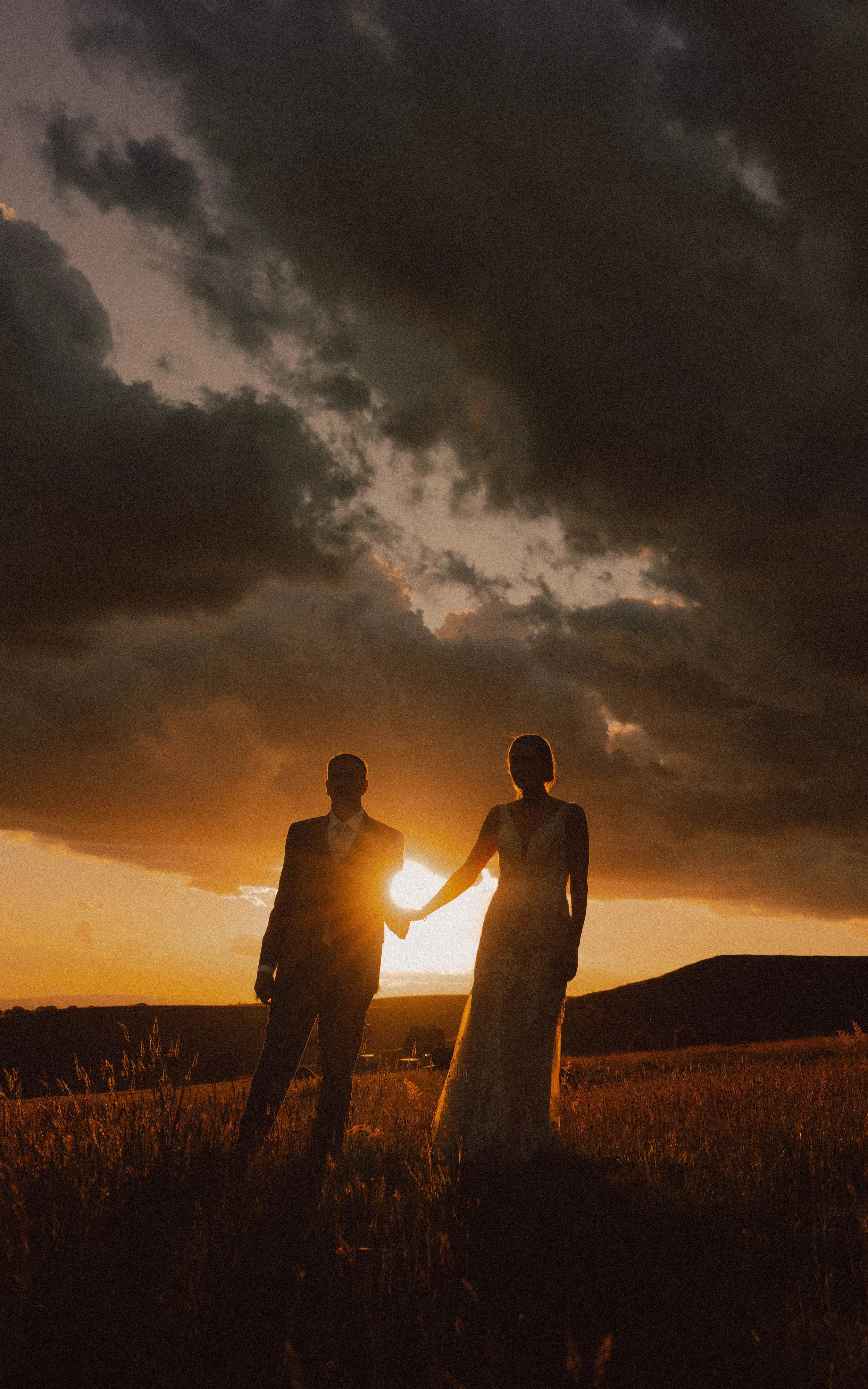 A couple in formal wedding attire holding hands at sunset with a dark, cloud-filled sky. silhouetted against the glowing sky.