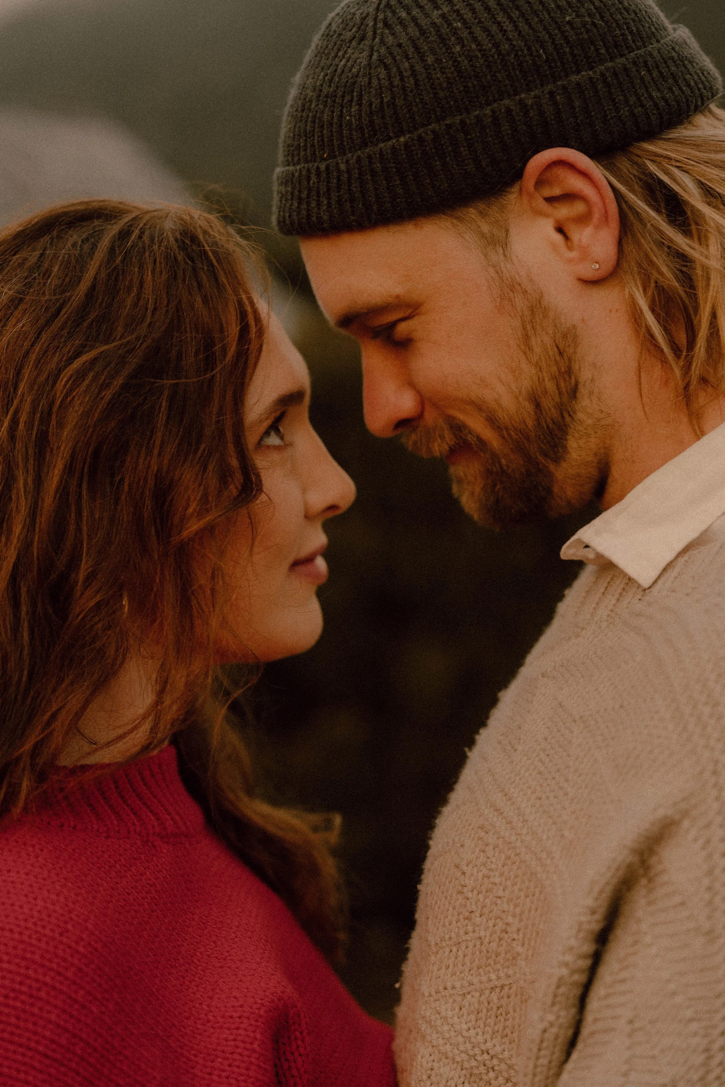 A couple with noses touching, eyes closed, sharing a close moment. The woman has long red hair and is wearing a red sweater. The man has shoulder-length blond hair, a beard, a black beanie, and a cream sweater.