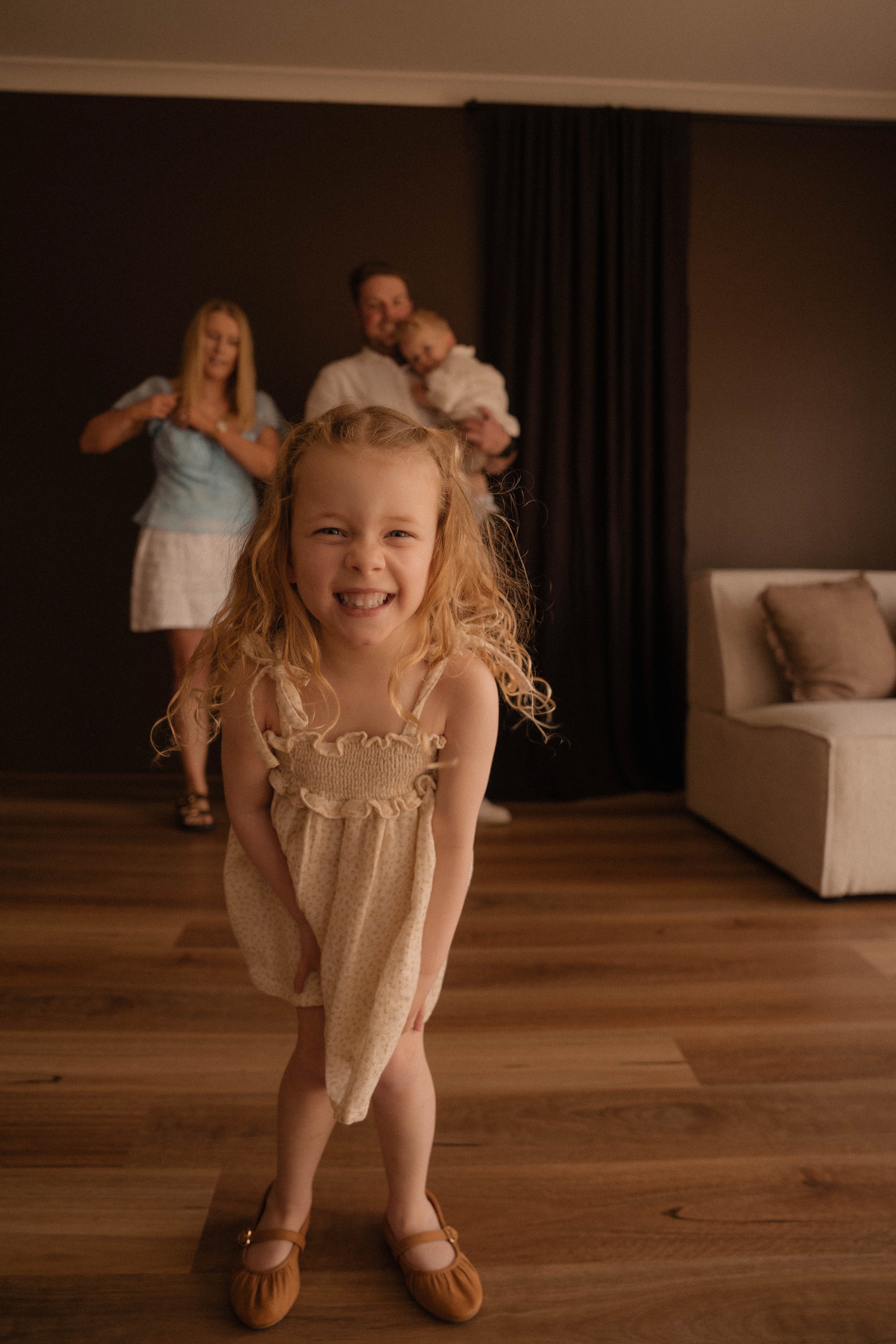 Young girl with curly blonde hair smiling at the camera in the foreground, three people in the background standing and smiling, inside a room with dark walls, curtains, and wooden flooring.