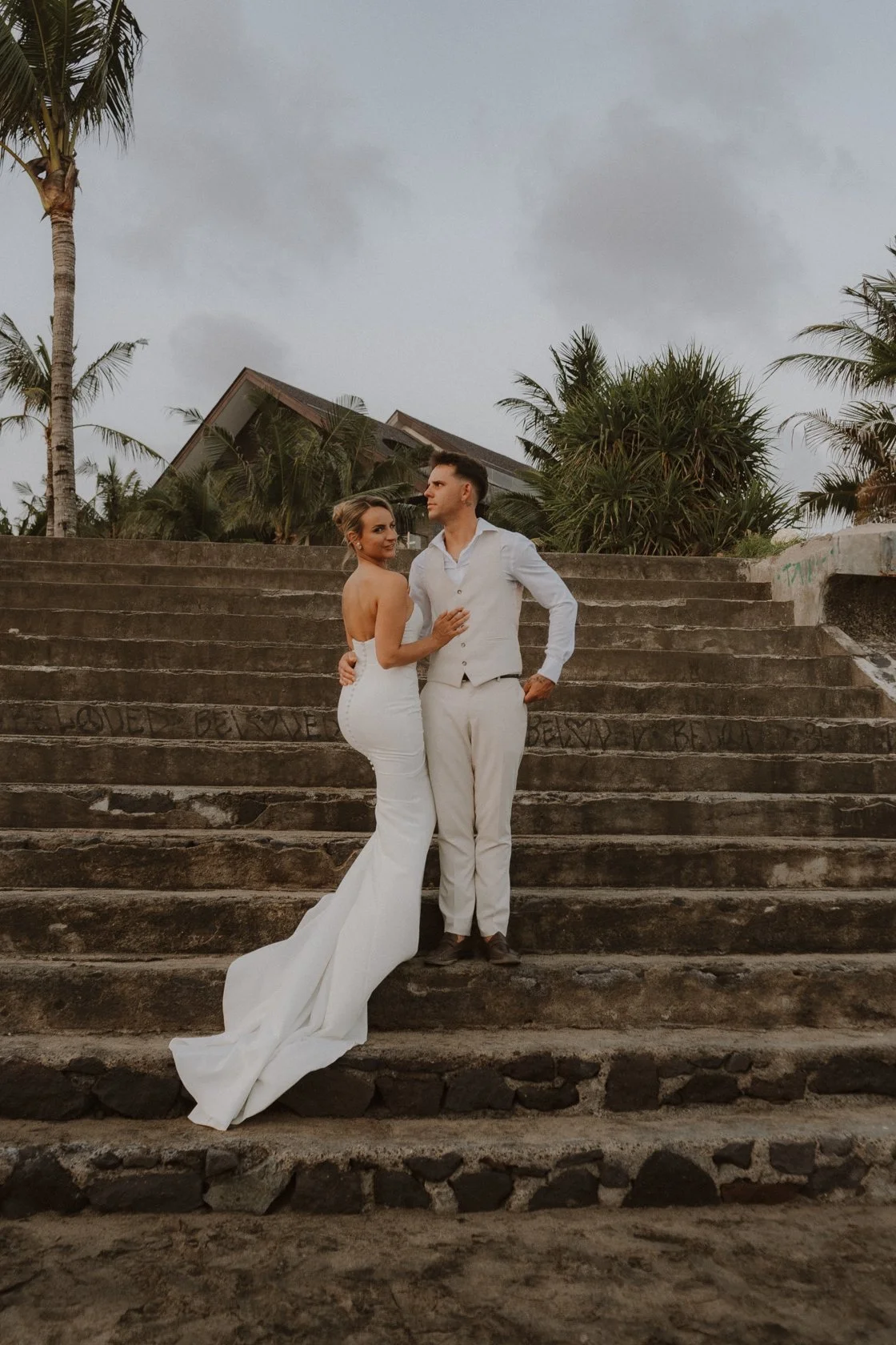 A bride and groom standing on stone stairs outdoors, with tropical trees and a house roof in the background, under cloudy skies. yarra valley and Melbourne wedding photographer