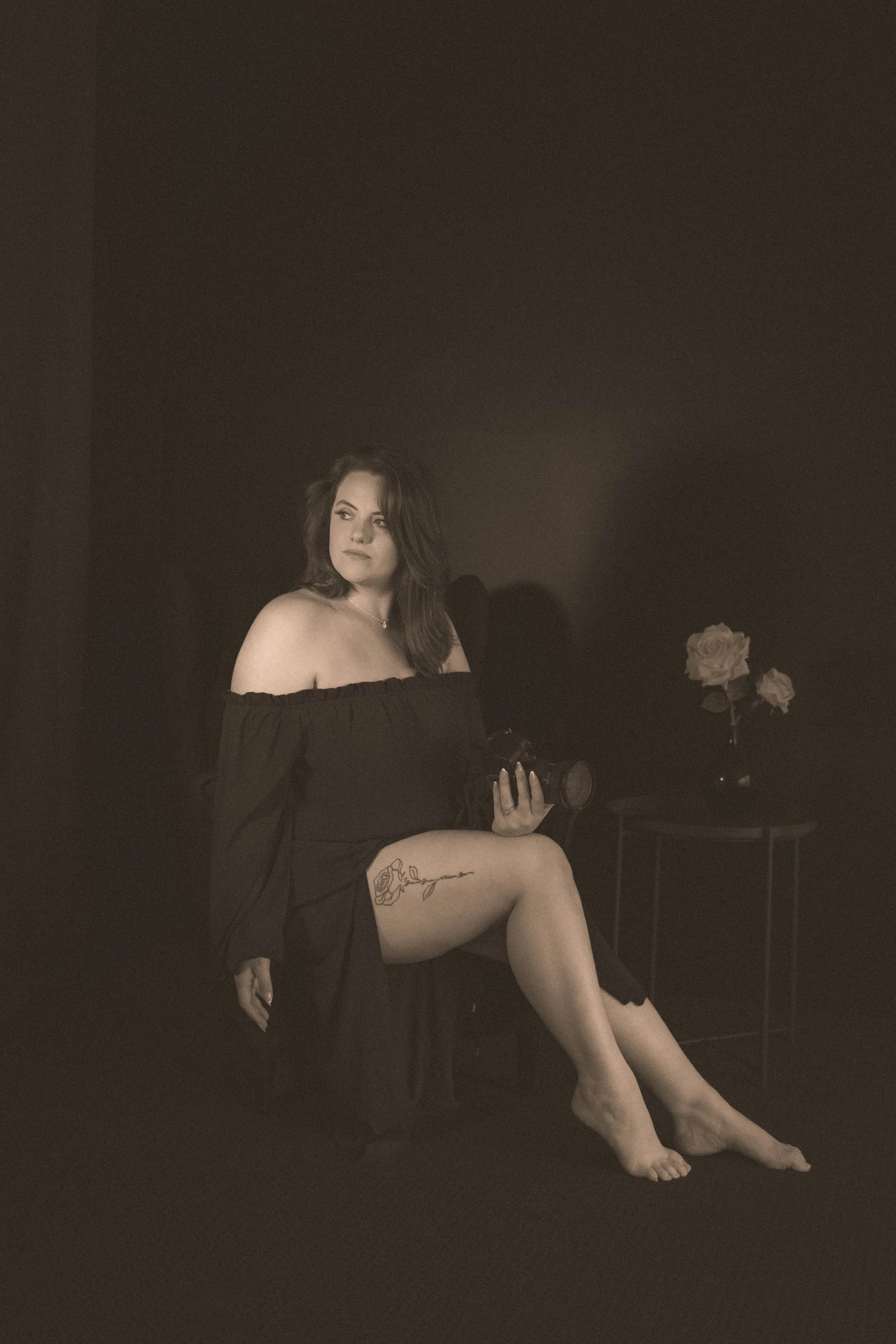 A woman sitting on a chair in a dark room creating a moody vibe, holding a camera, with roses on a table beside her, wearing a black off-the-shoulder dress, showing a tattoo of a rose on her thigh. branding images for jess at and photography
