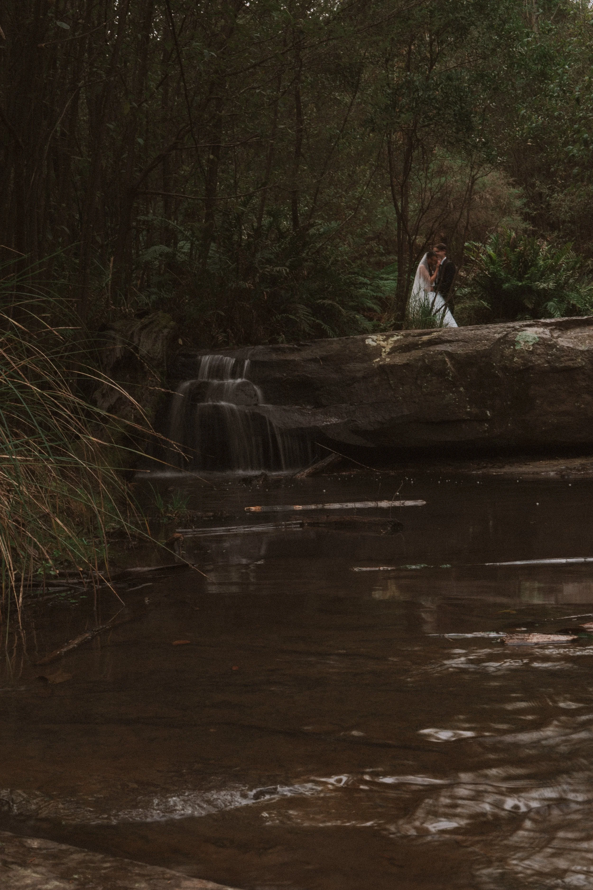 A couple in wedding attire stands on a large rock beside a small waterfall in a dense forest, embracing each other.
