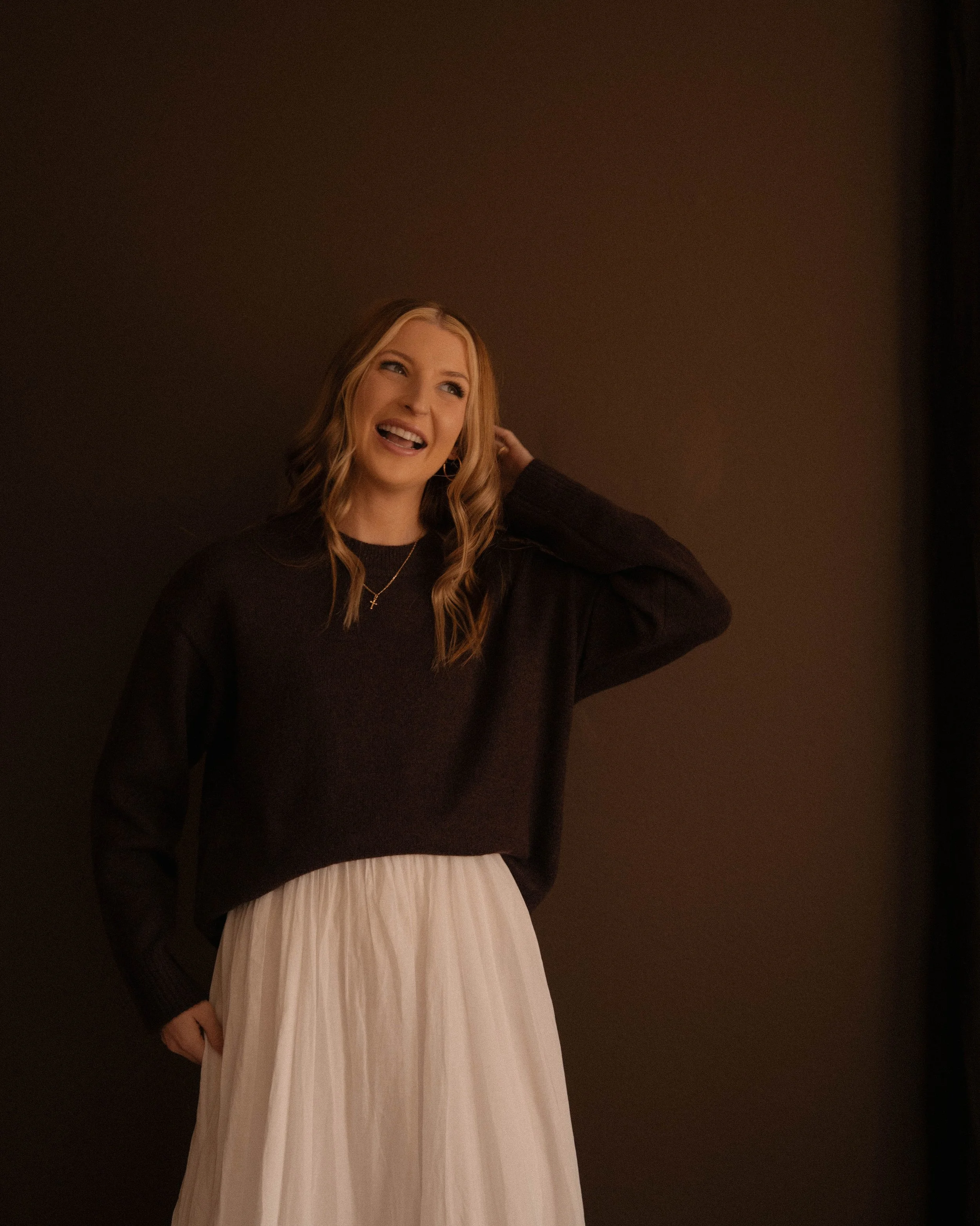 A woman with wavy blonde hair, wearing a black sweater with a cross necklace, smiling and touching her hair, standing against a dark background.