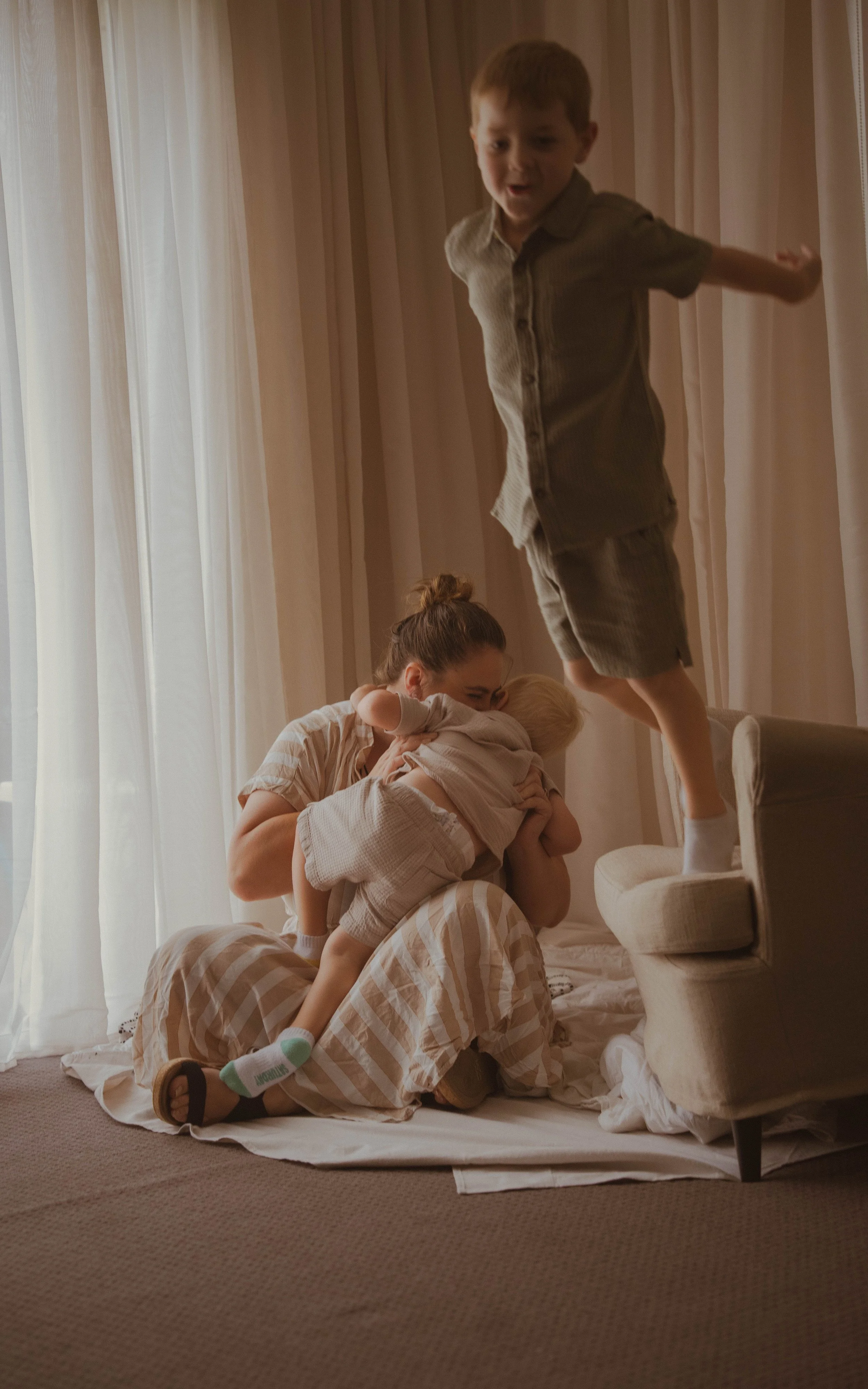 A woman sitting on the floor with a baby in her lap, holding the child close to her face, while a young boy jumps from a chair behind them, all indoors with light curtains in the background.