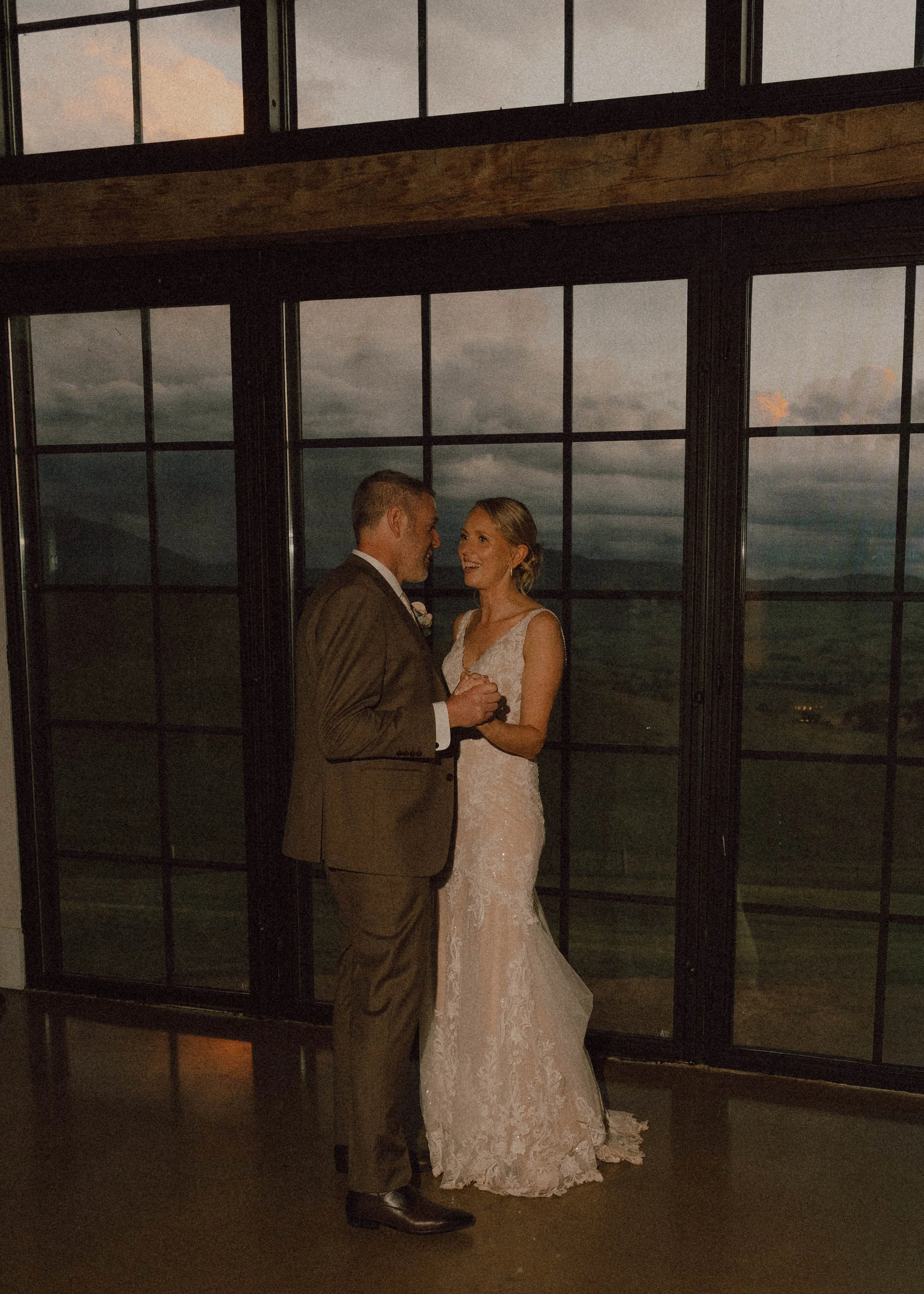 A bride and groom sharing a dance at their wedding reception, standing in front of large windowpanes with a sunset view.