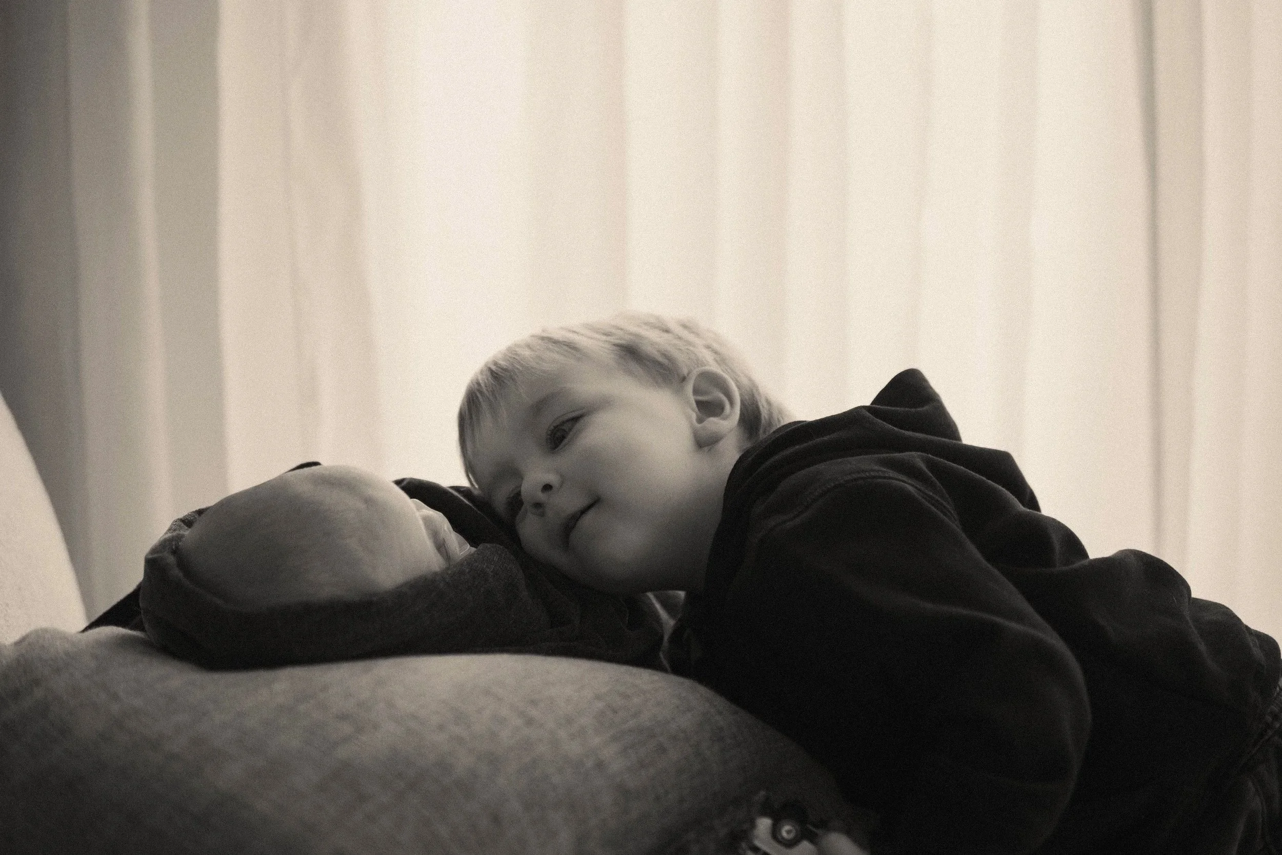 A young boy  with blonde hair lying on a couch, smiling and looking at a his new born little brother with so much love