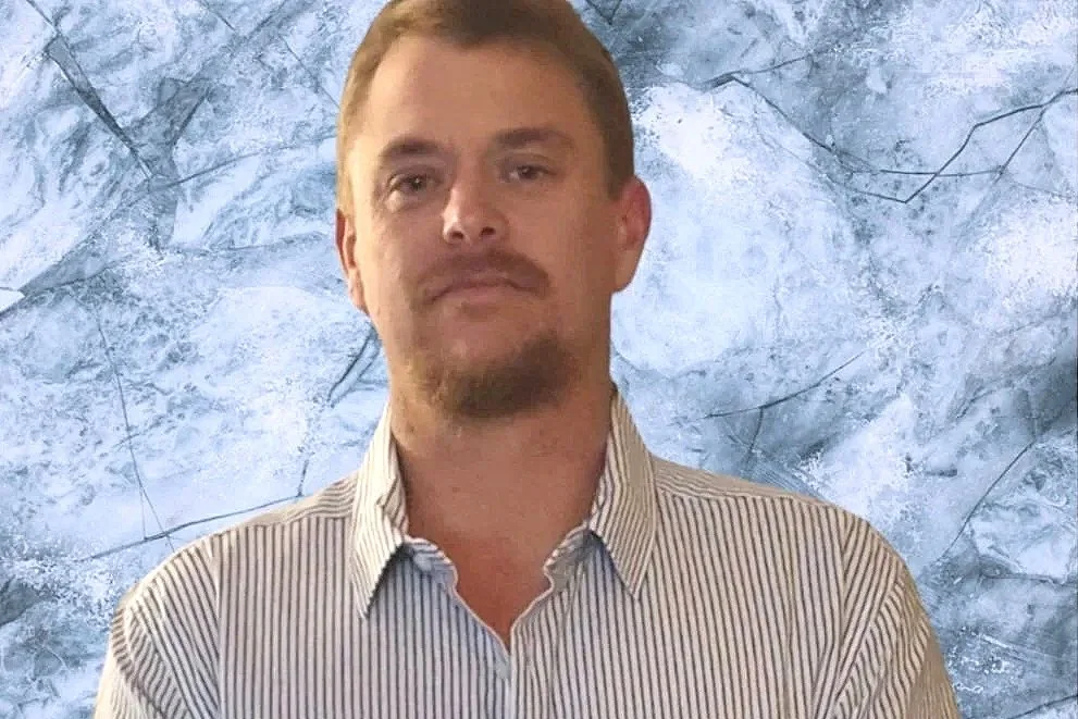 A man with light brown hair, a short beard, and wearing a striped shirt stands against a textured blue background.