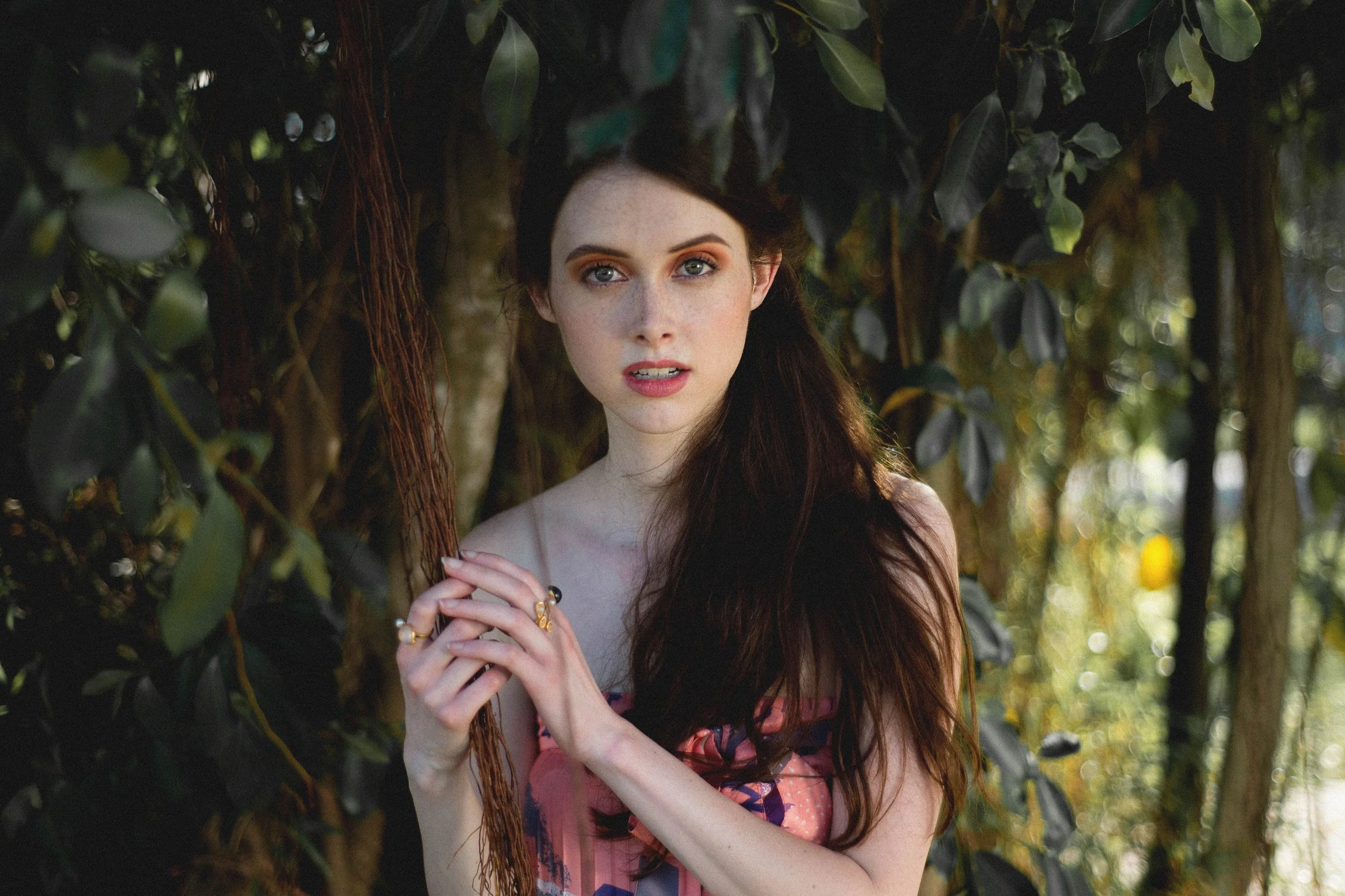 A young woman with red hair and blue eyes standing outdoors among trees and greenery, looking directly at the camera with a neutral expression.