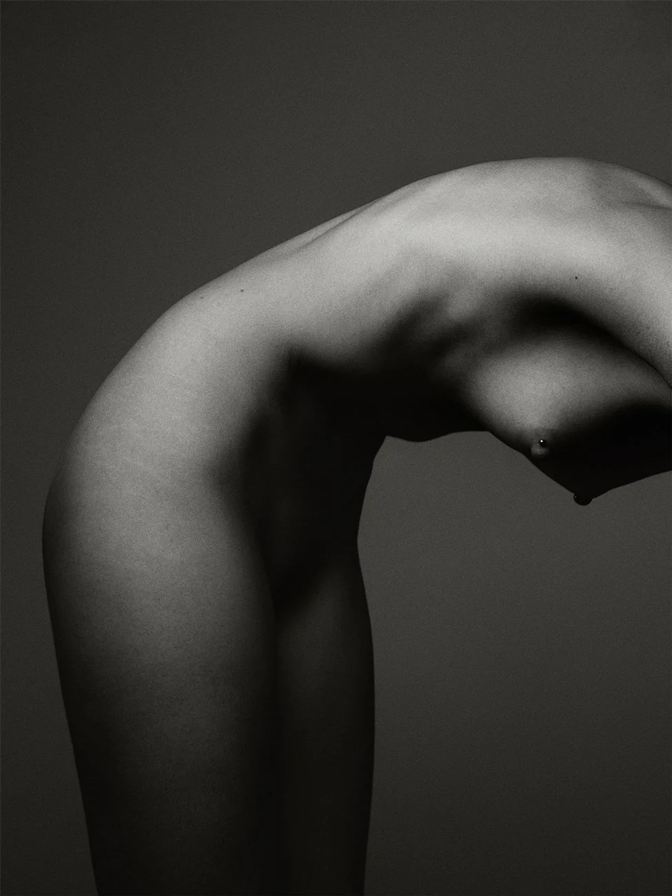 Black and white photograph of a person's back and arm, arching and leaning forward, with a small piercing visible on the side of the chest.