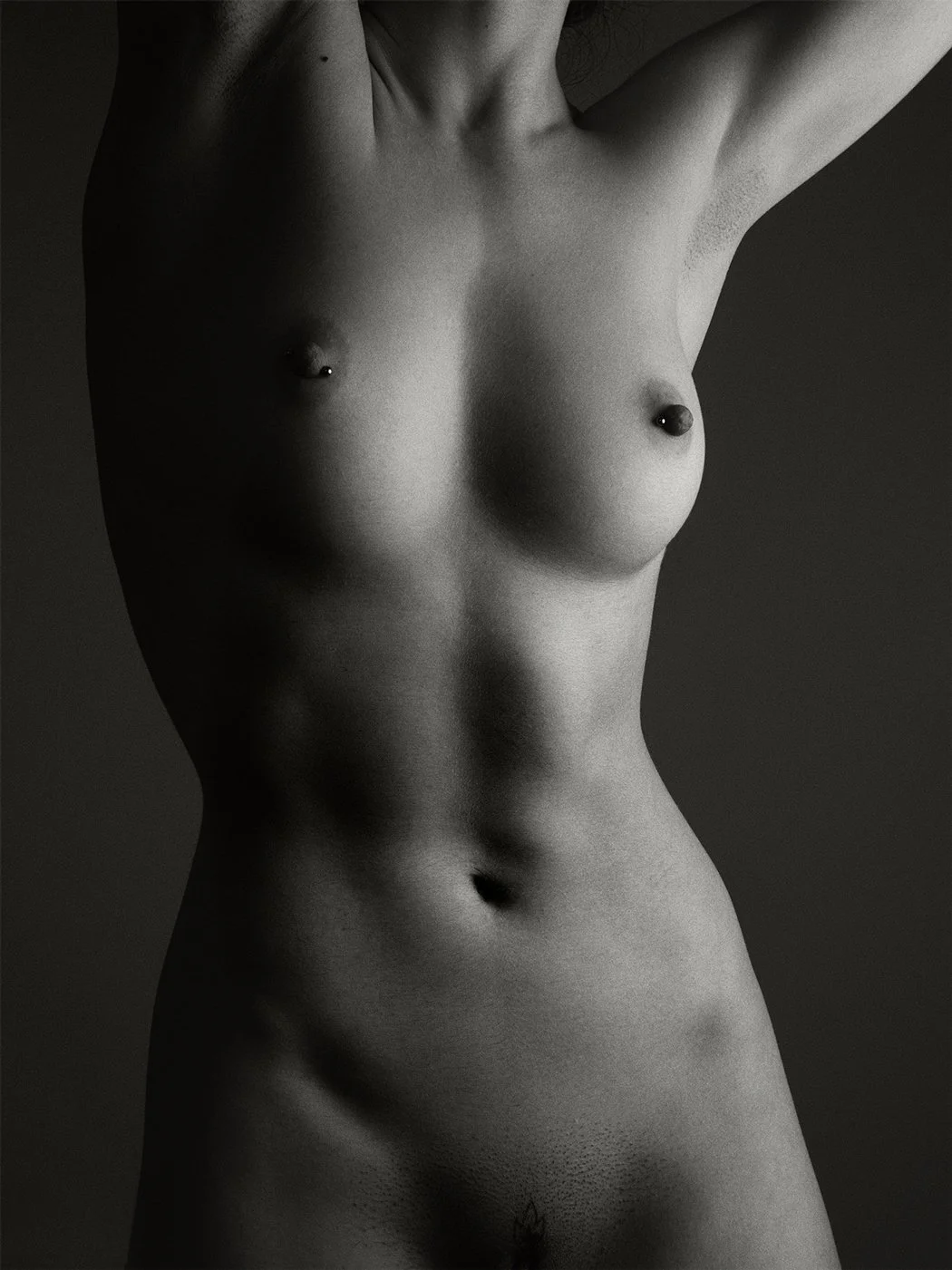 Black and white photo of a nude female torso with arms raised, showing part of her chest and abdomen.
