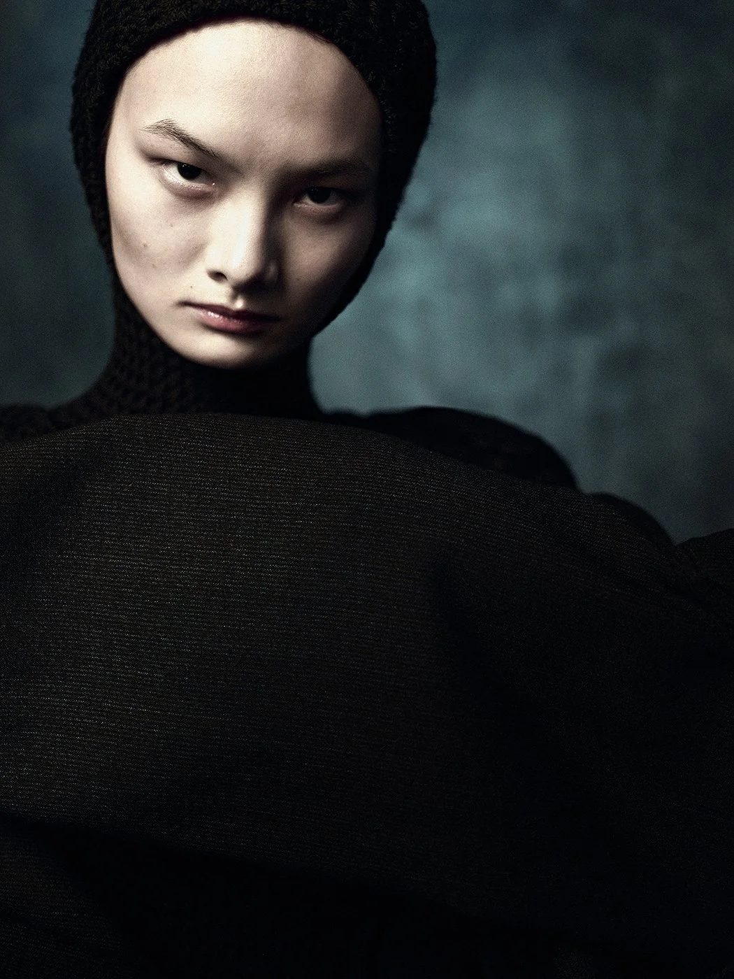 A woman with fair skin and short dark hair, wearing a black textured turtleneck and a matching head covering, poses against a dark background with a serious expression.