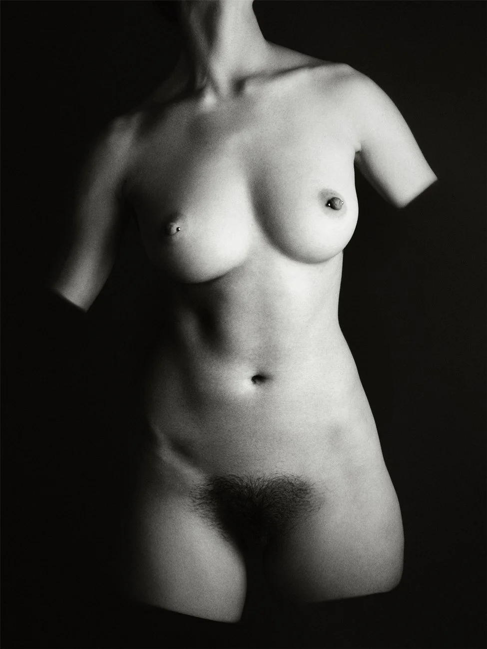 Black and white photograph of a nude woman, showing her torso and upper thighs, with tattoos on her chest.