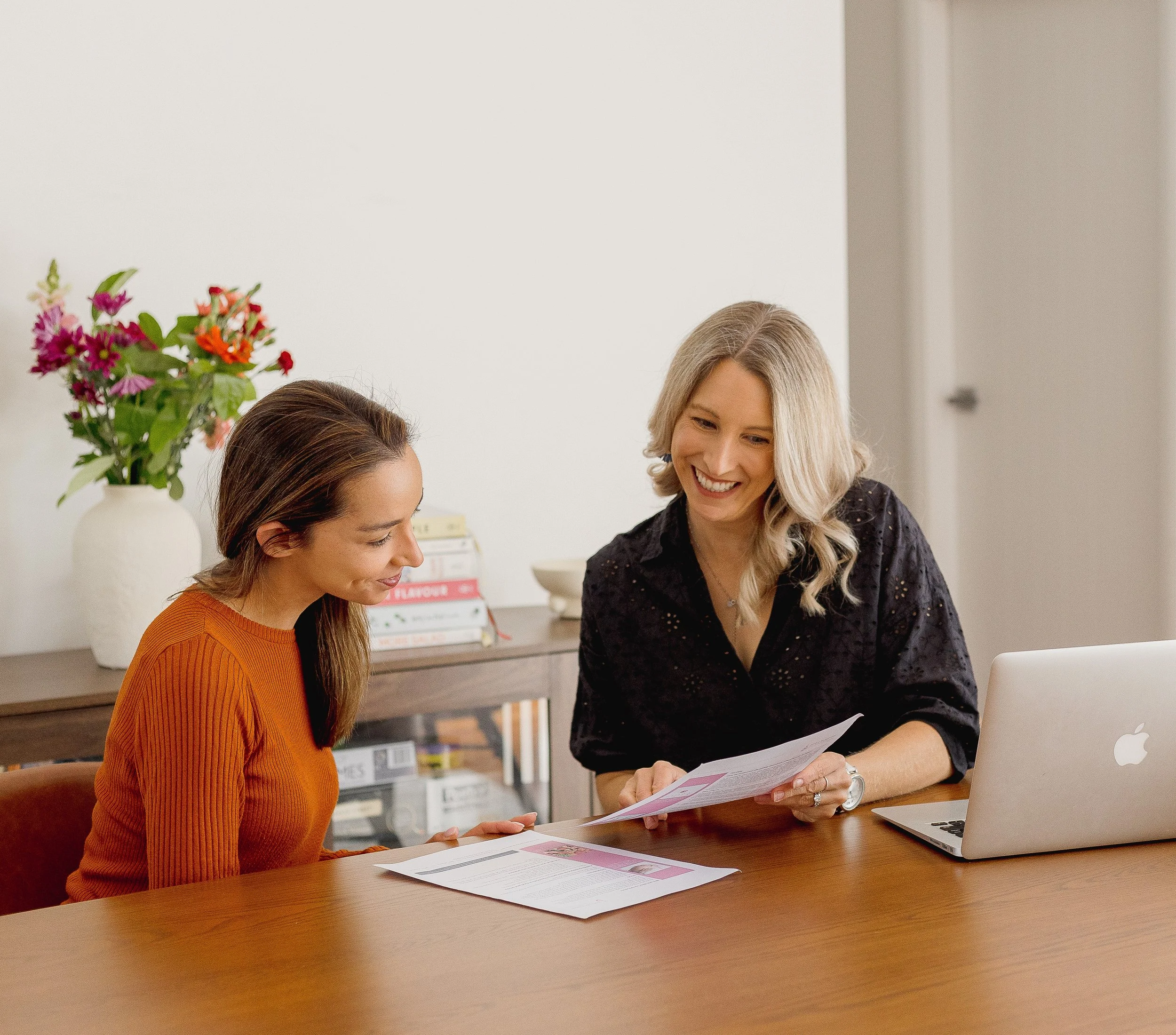 Two women sitting at a wooden table having a discussion, with papers and a laptop in front of them. One woman is wearing an orange sweater, and the other is wearing a black top. Behind them is a bookshelf with books and a large white vase with colorful flowers.