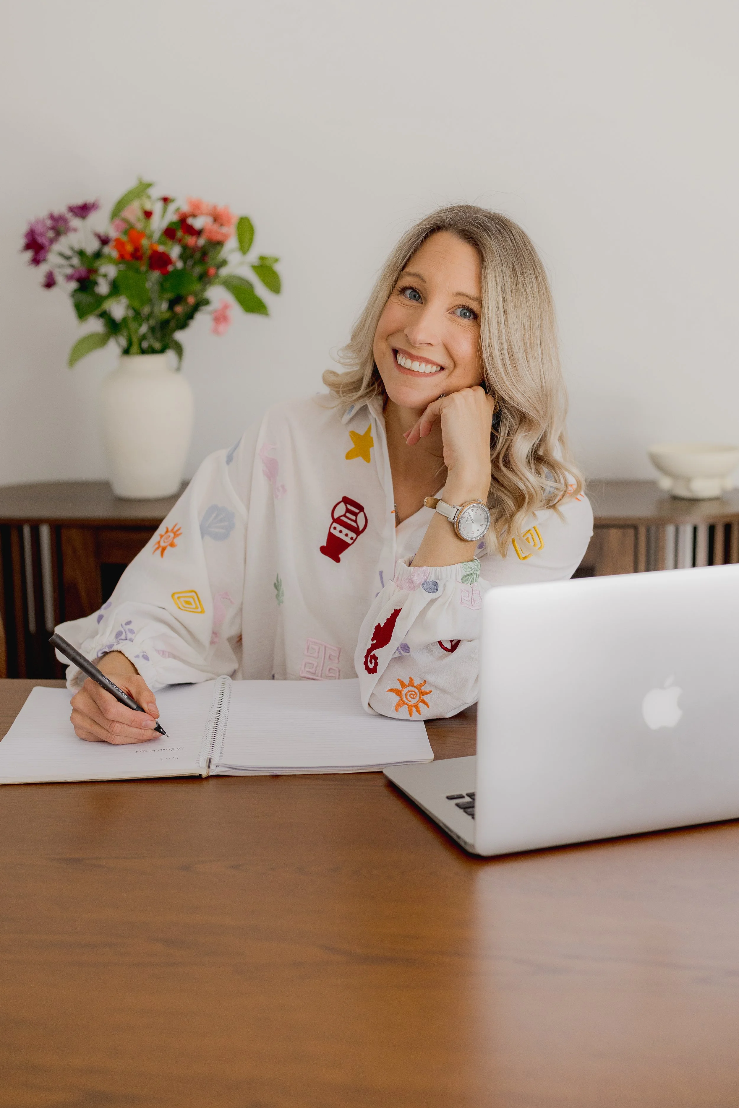 A woman with blonde hair, smiling, sitting at a desk with a laptop and a notebook, wearing a white shirt with colorful embroidered patterns, in a bright room with a vase of flowers in the background.