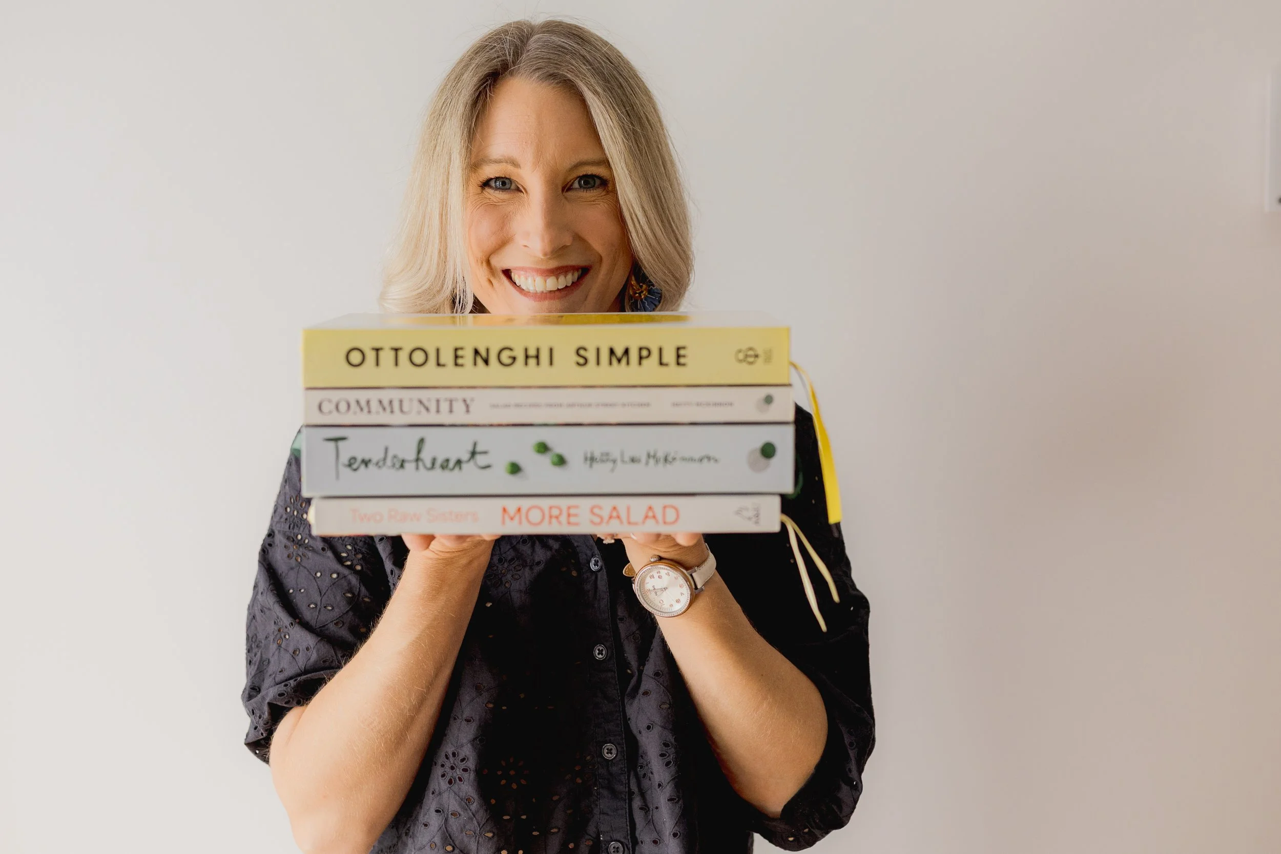 A woman with blonde hair holding a stack of five books, smiling, wearing a black eyelet blouse and a watch on her left wrist, standing against a plain white background.